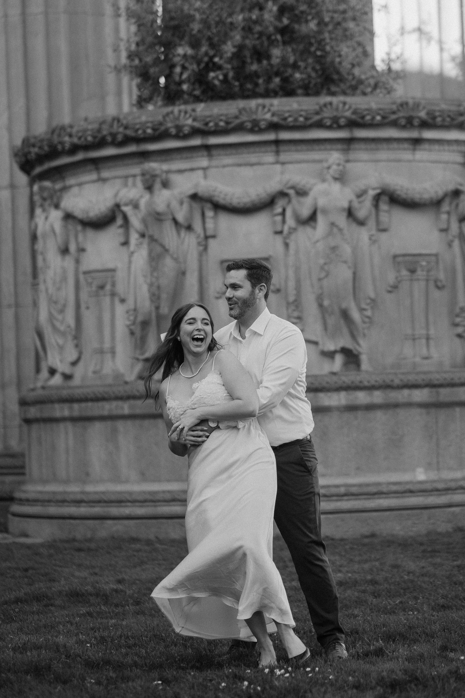 Joyful couple laughing and dancing in front of an ornate stone fountain during engagement photos.