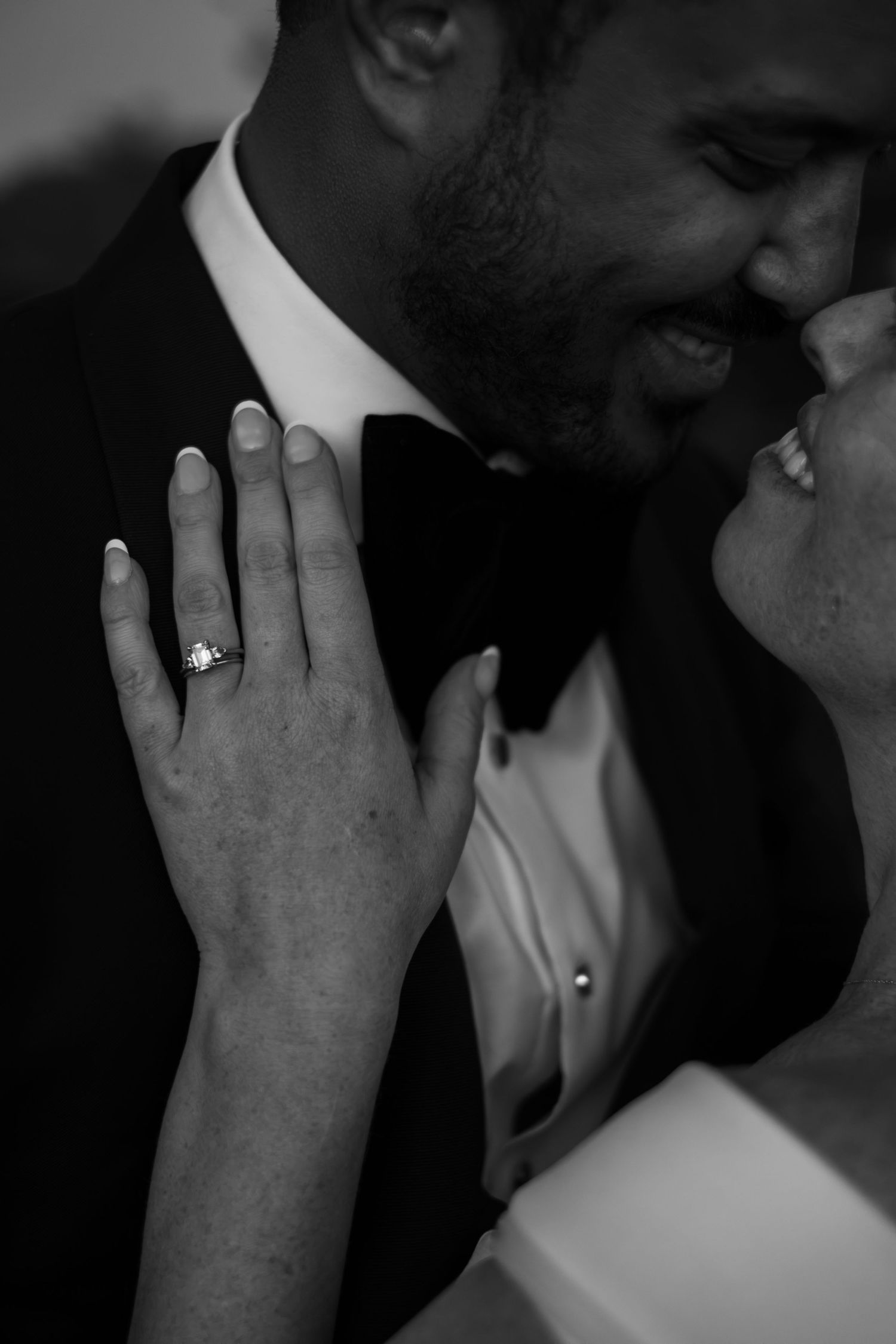Black and white photo of a couple in formal wear almost kissing, bride showcasing diamond engagement ring on groom's chest.