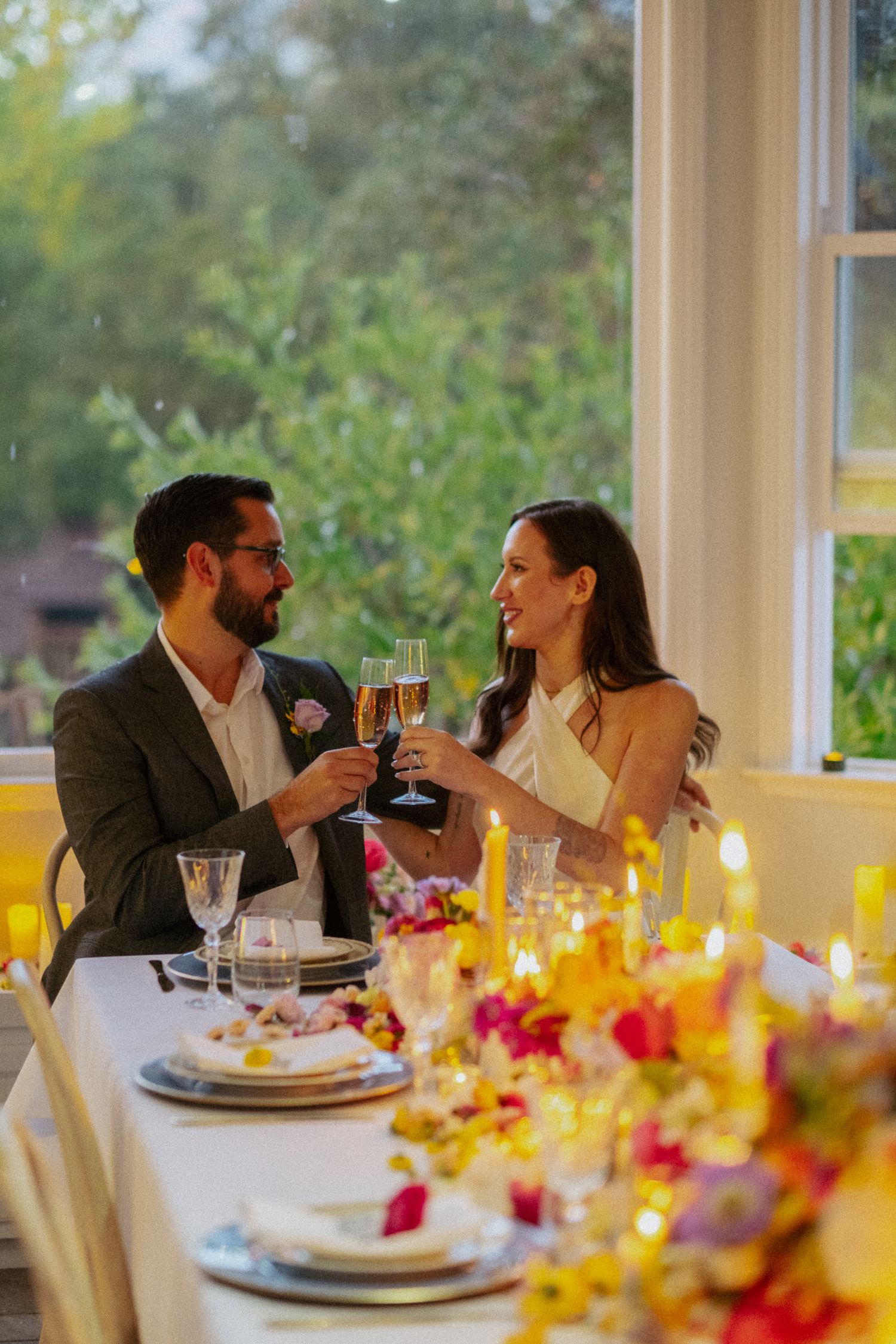 Bride and groom in elegant attire clink champagne glasses over a floral-decorated wedding reception table by bright windows.