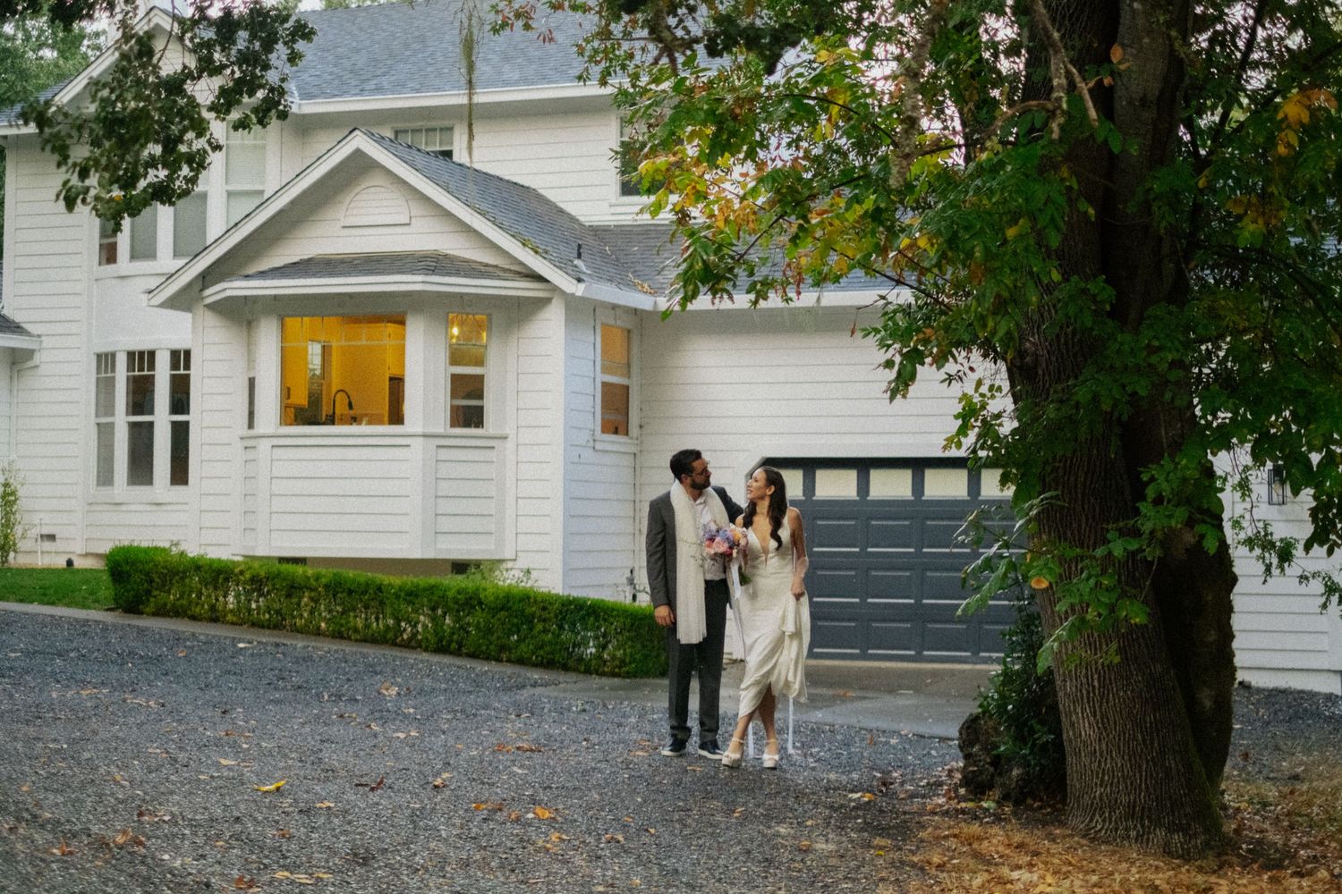 Couple walking in front of a white farmhouse with a garage, surrounded by trees on a gravel driveway.