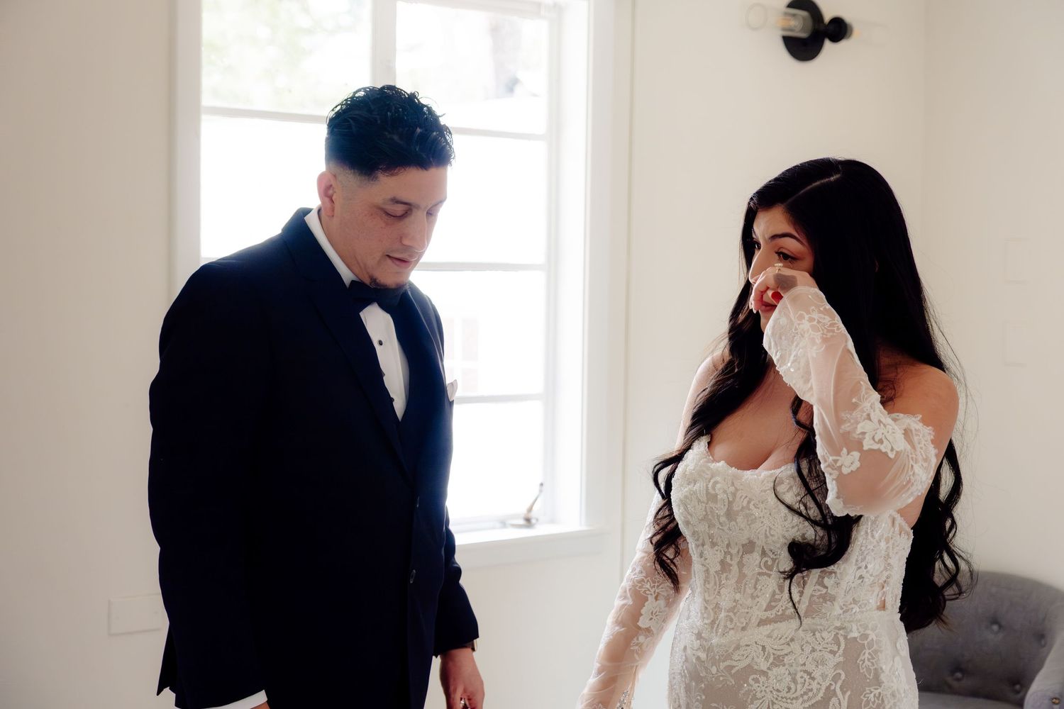 Emotional bride in lace wedding gown wiping tears beside groom in black tuxedo during wedding day first look.