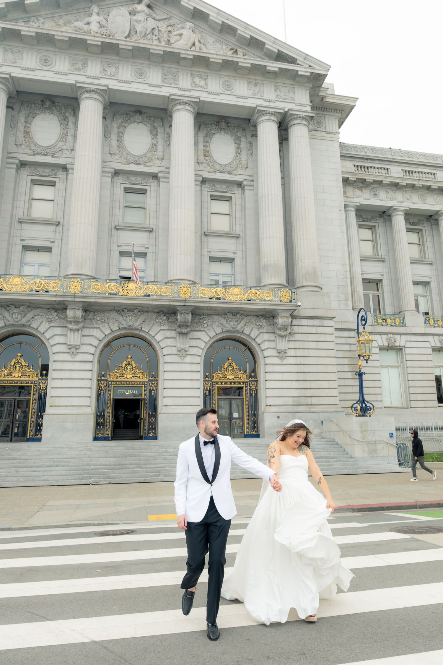 Bride and groom walk hand-in-hand across a crosswalk in front of San Francisco City Hall.
