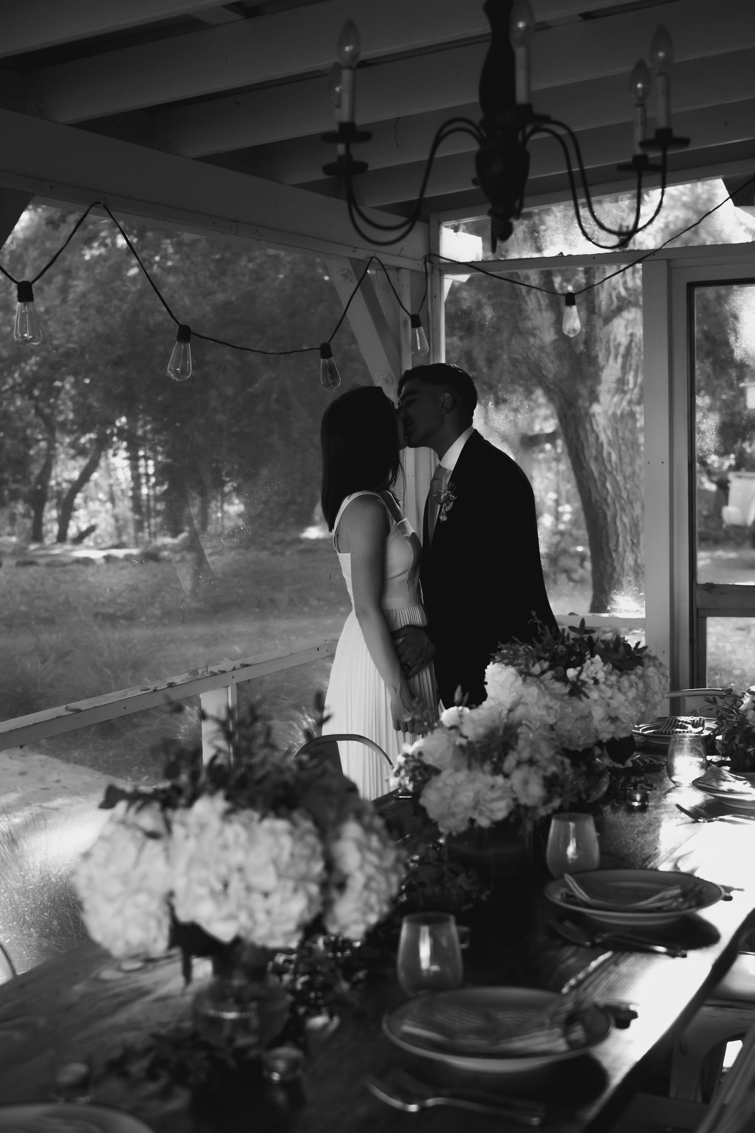 Black and white photo of a couple sharing a kiss at their wedding reception table adorned with floral arrangements.