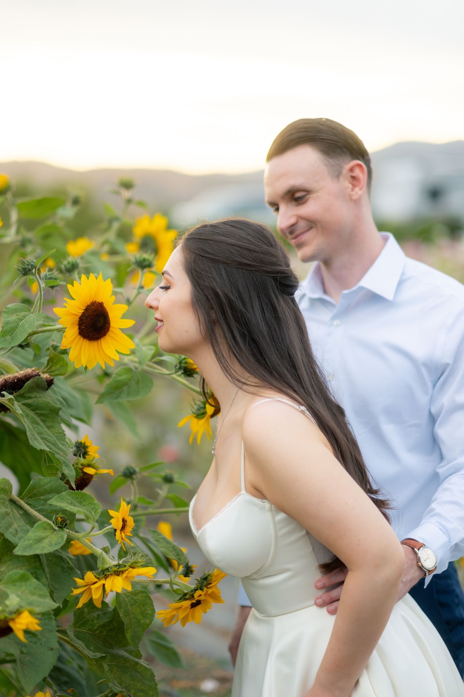 Couple embracing near vibrant sunflowers at sunset, bride smelling blooms as groom smiles lovingly behind her.