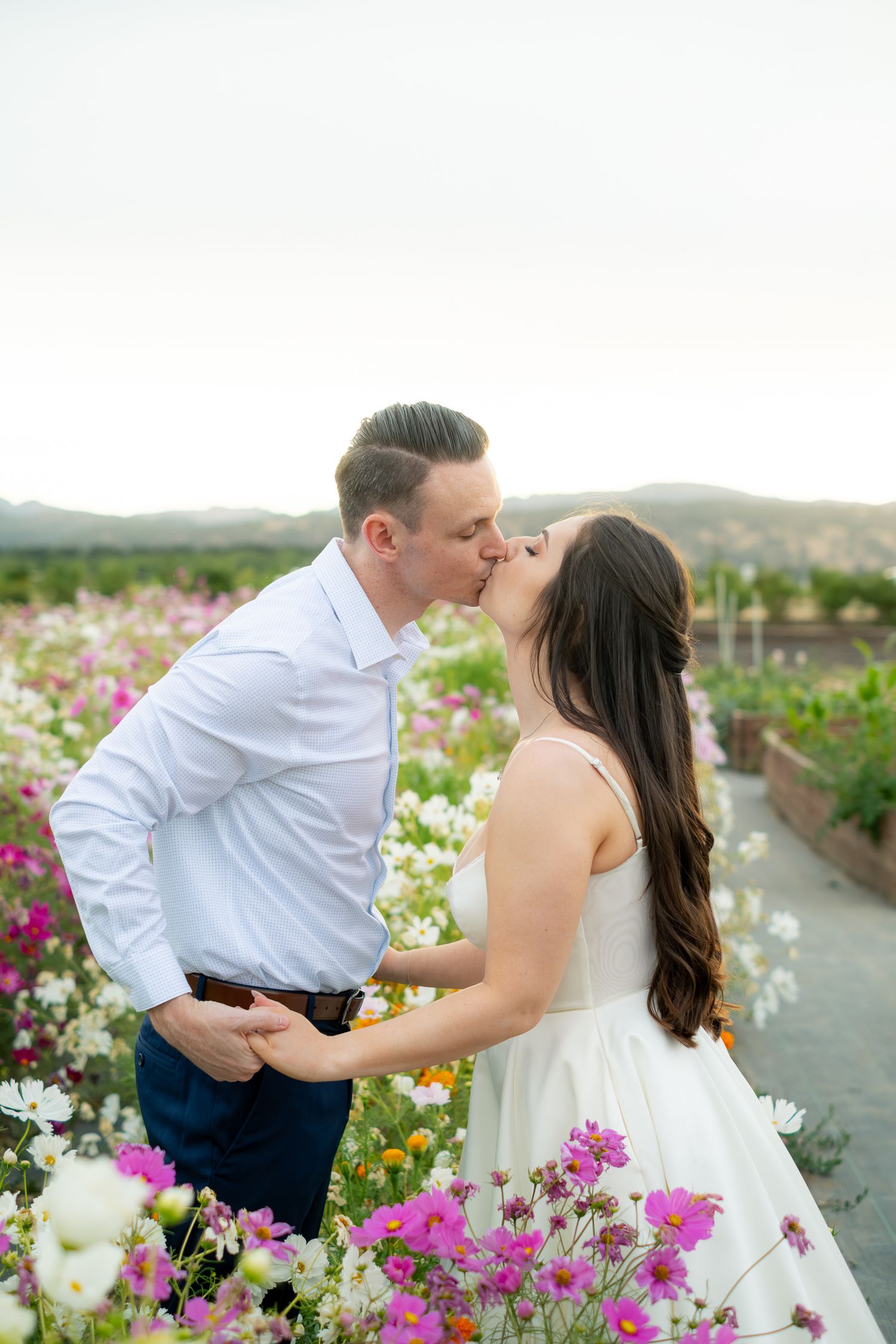 Couple sharing a romantic kiss in a vibrant flower field with colorful blooms and mountain views in the background.