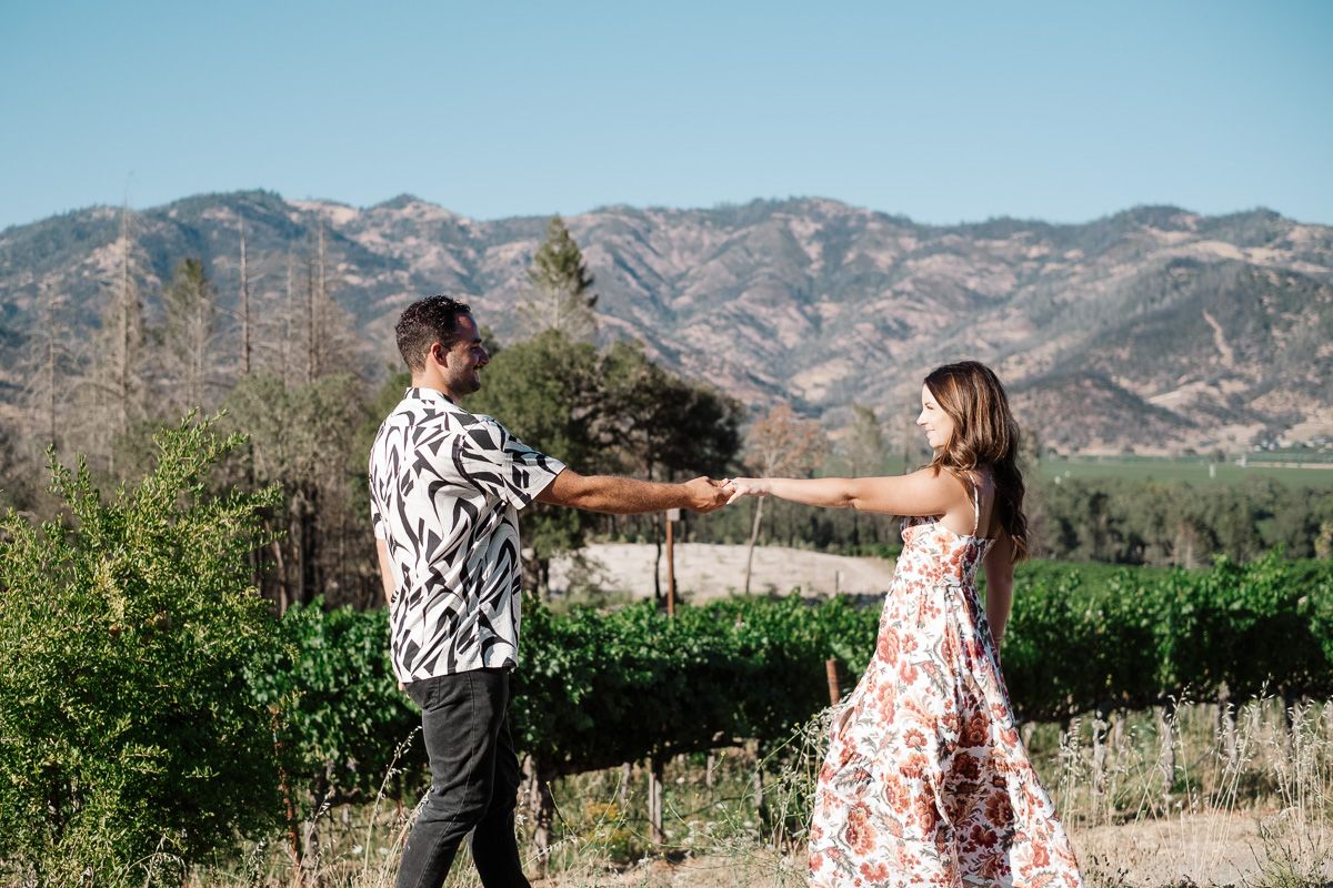 Couple holding hands walking through a vineyard with mountain views in Napa Valley.