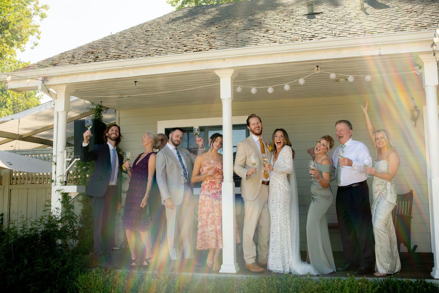 Wedding party poses and celebrates on white porch with string lights in sunny outdoor setting.