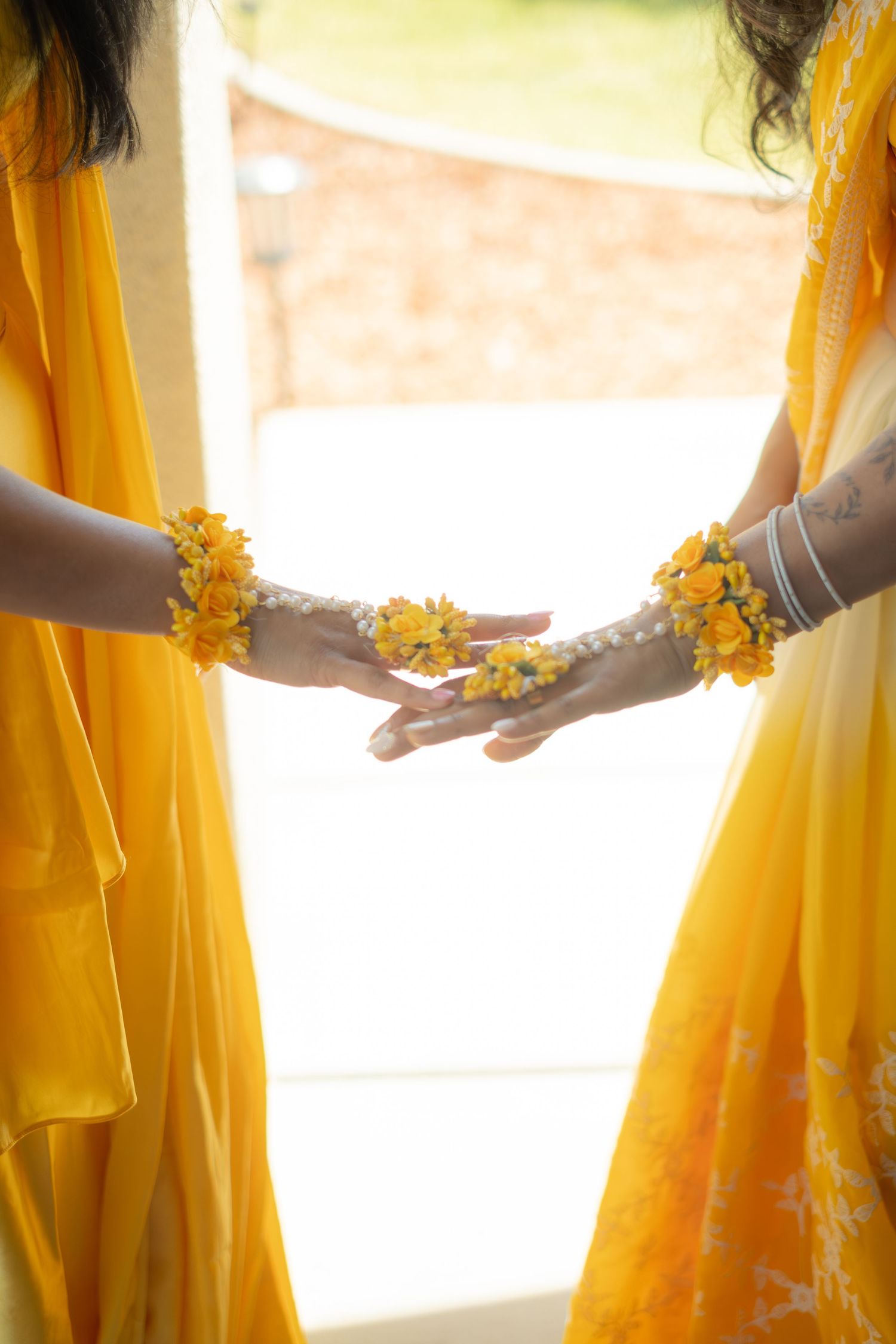 Two women in yellow sarees hold hands adorned with floral bracelets and henna at an Indian wedding ceremony.