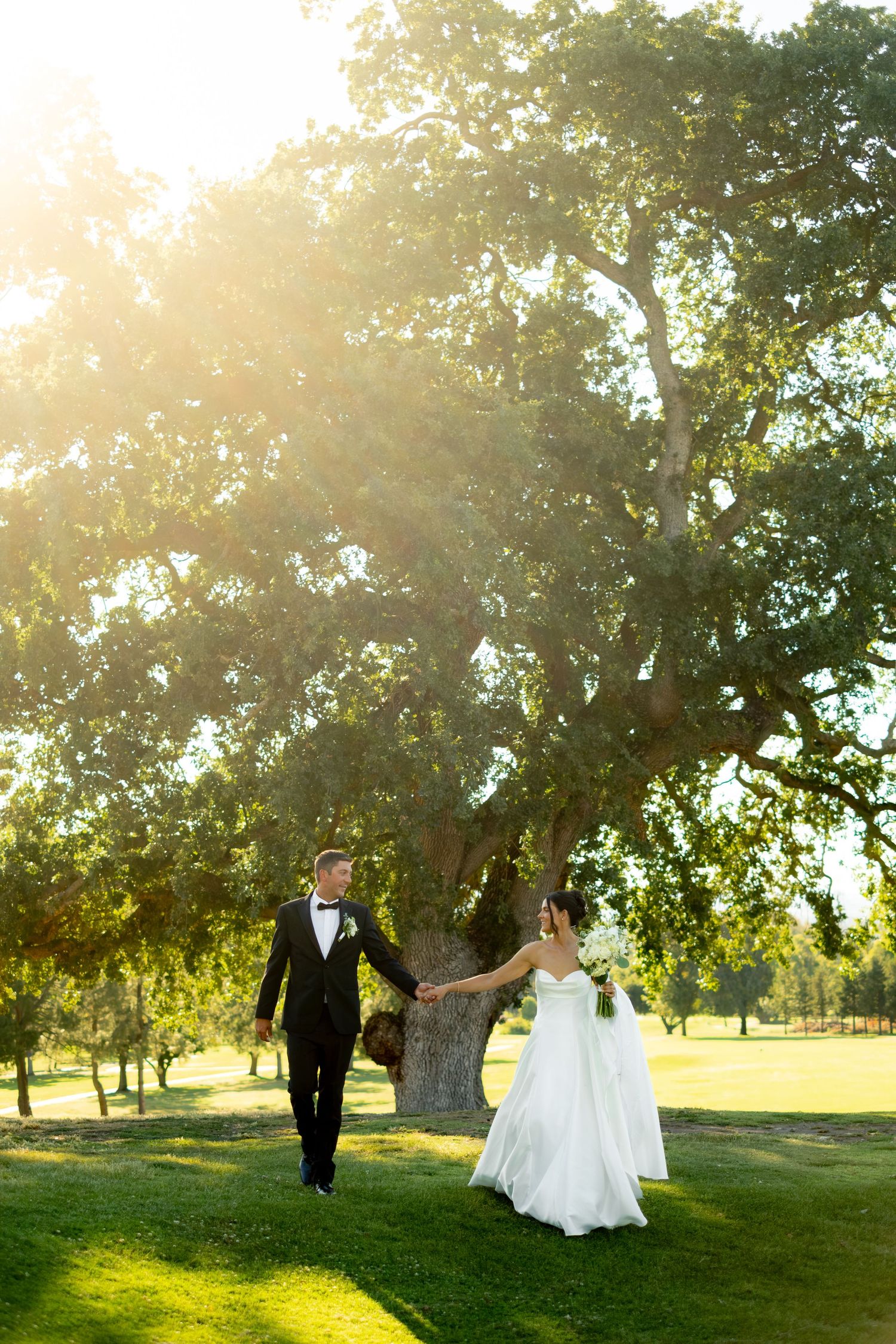 Bride and groom holding hands walking under large oak tree bathed in golden sunlight at outdoor wedding.