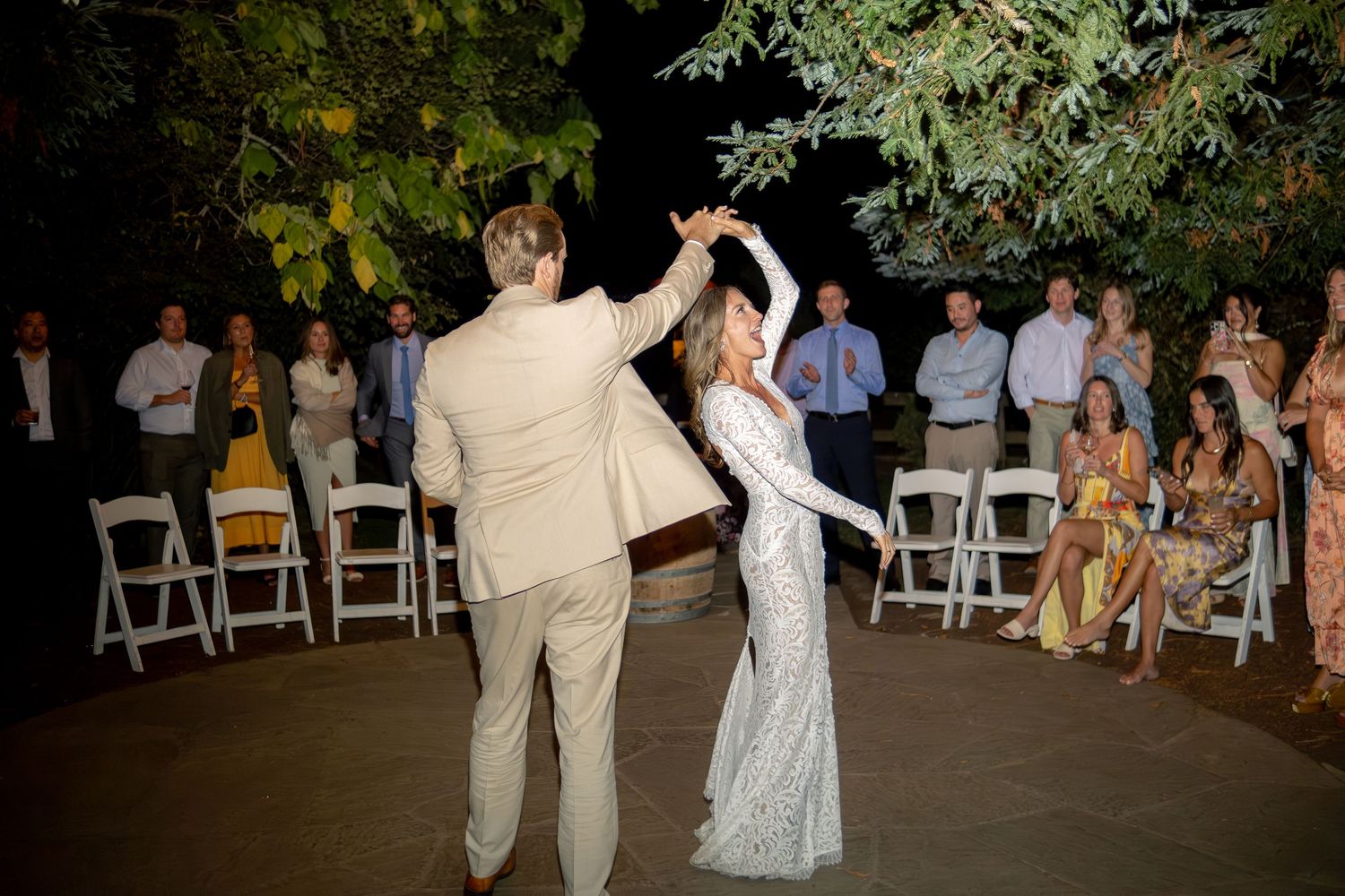 Groom in tan suit twirling bride in lace gown during first dance at outdoor wedding.