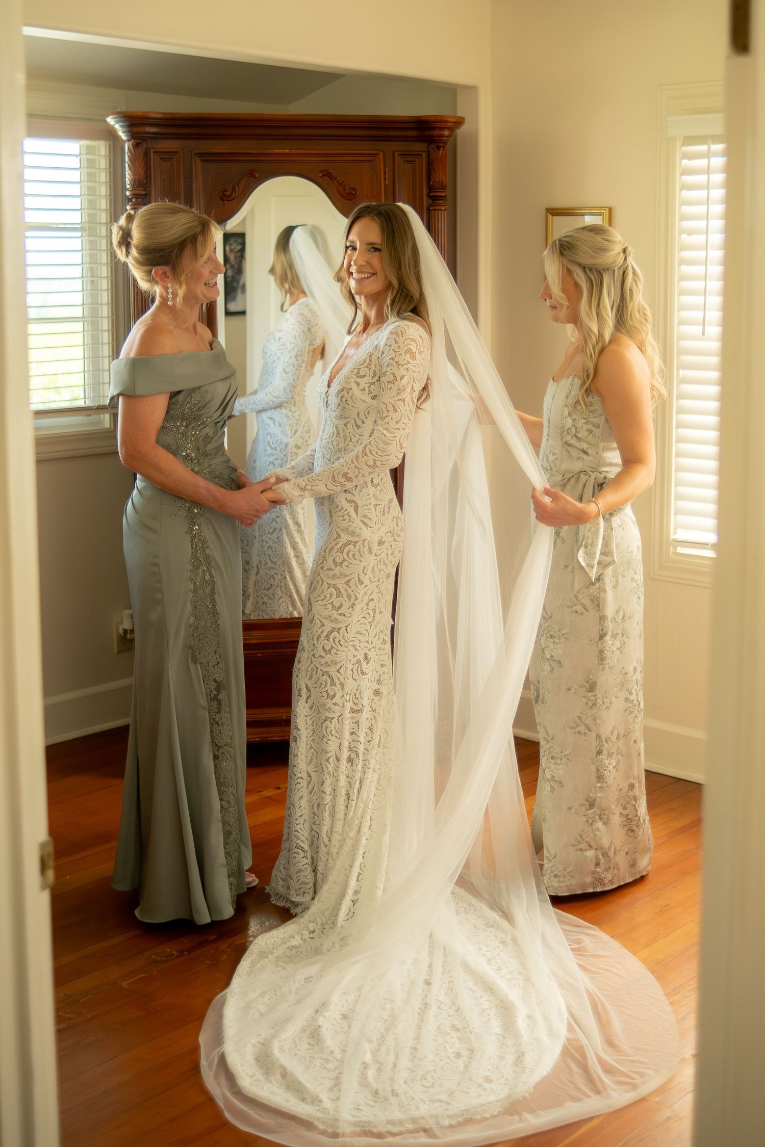 Bride in lace gown and veil stands with two bridesmaids in sage green dresses during wedding preparations.