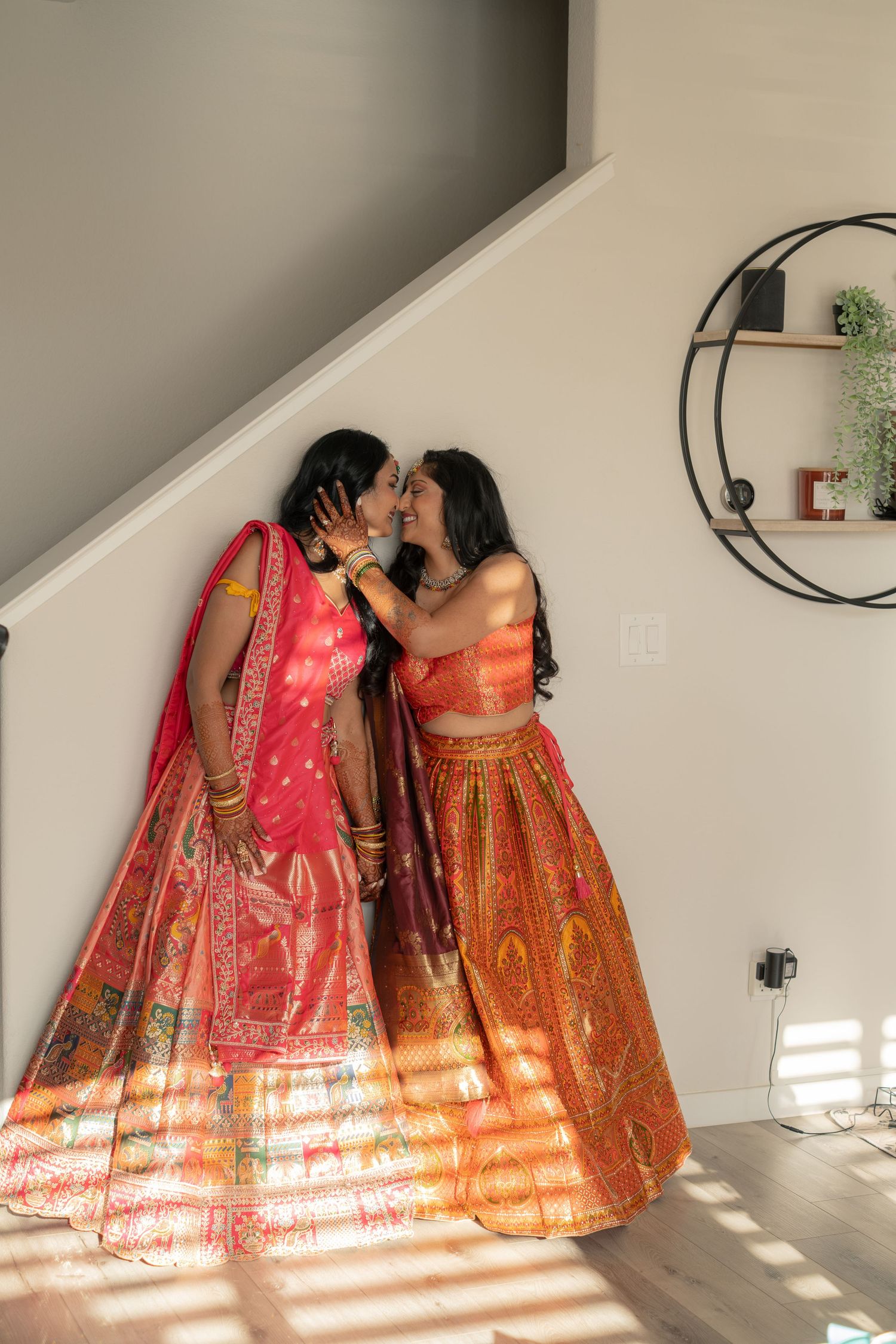 Two women in colorful Indian lehengas share a tender moment near a sunlit staircase.