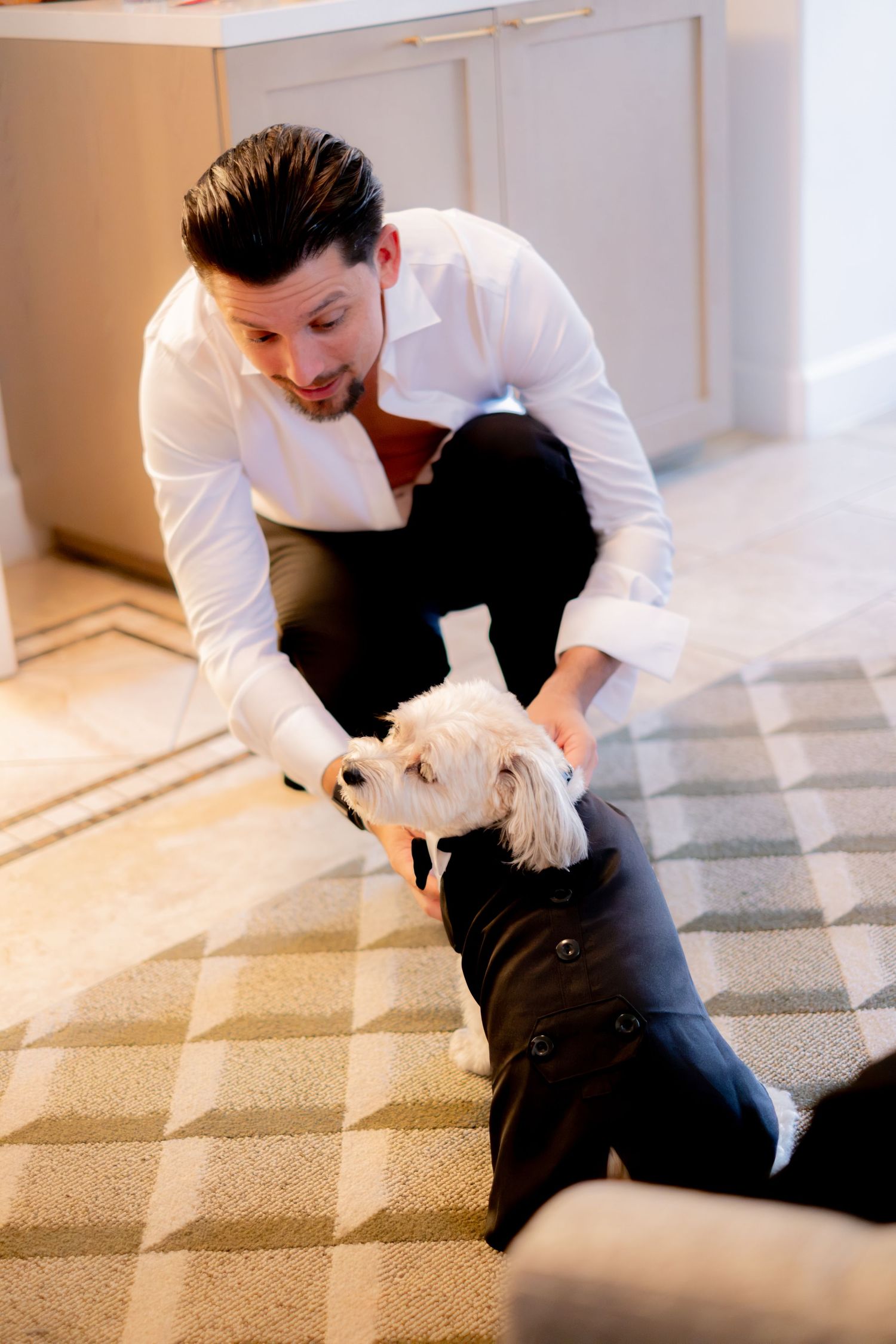 Groom in white dress shirt kneels on floor to pet small white dog dressed in tuxedo.