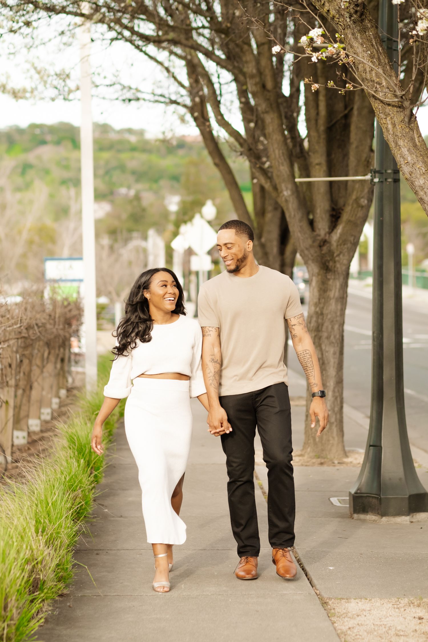Happy couple holding hands and walking along a sunny tree-lined sidewalk in a charming town.