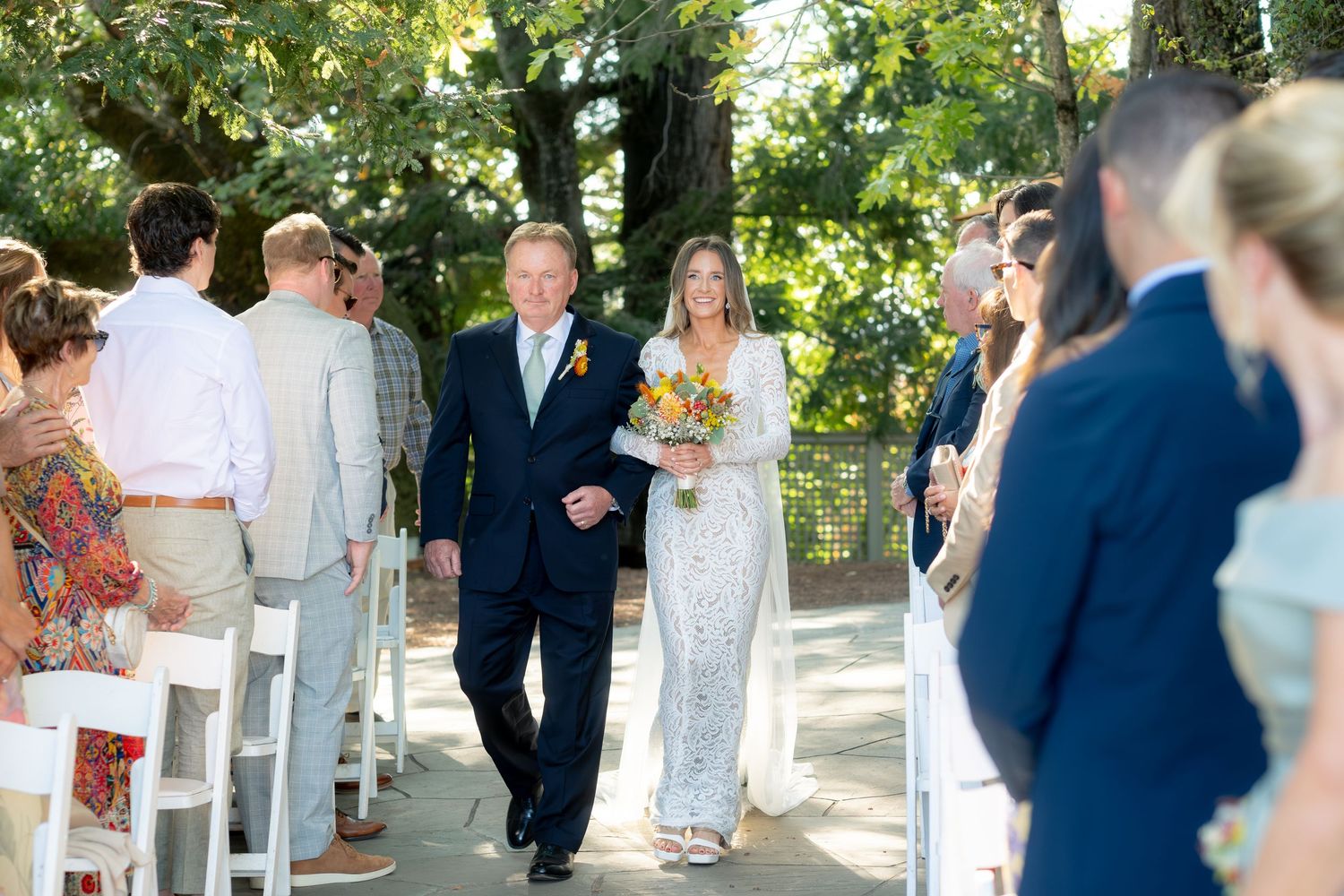 Bride in white lace gown walks down outdoor aisle with older gentleman at sunny garden wedding ceremony.
