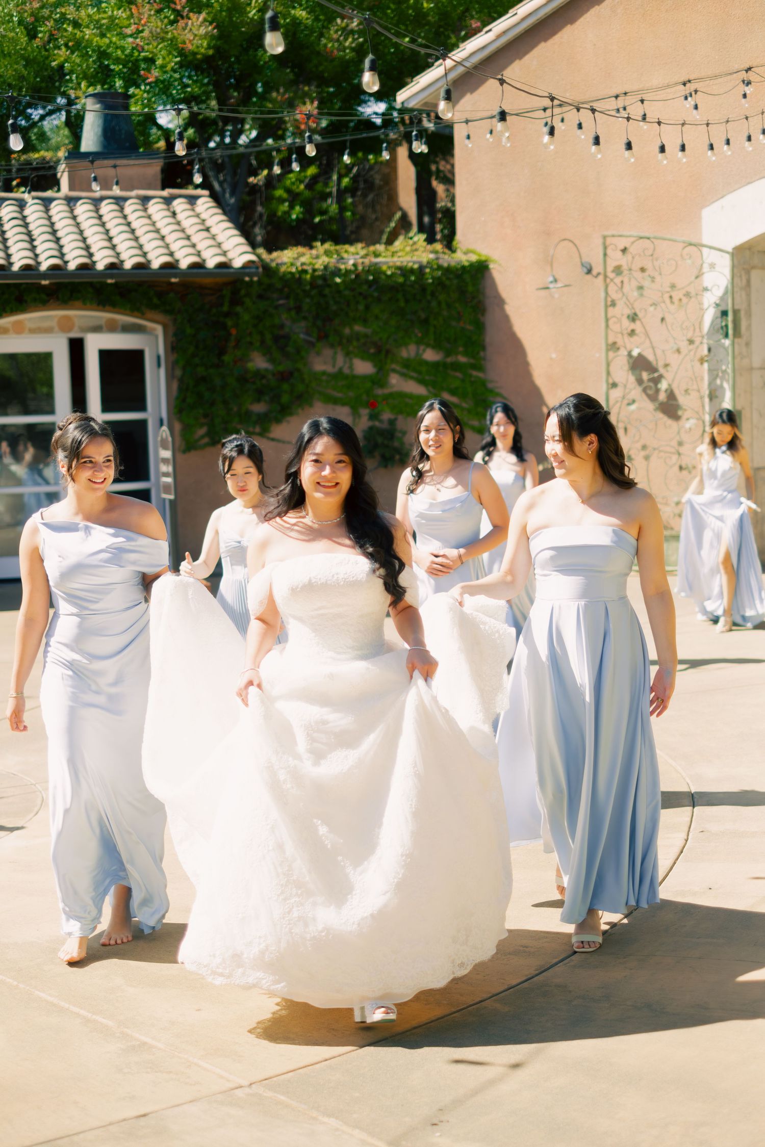 Bride in white gown walks with bridesmaids in light blue dresses outside a sunlit venue.