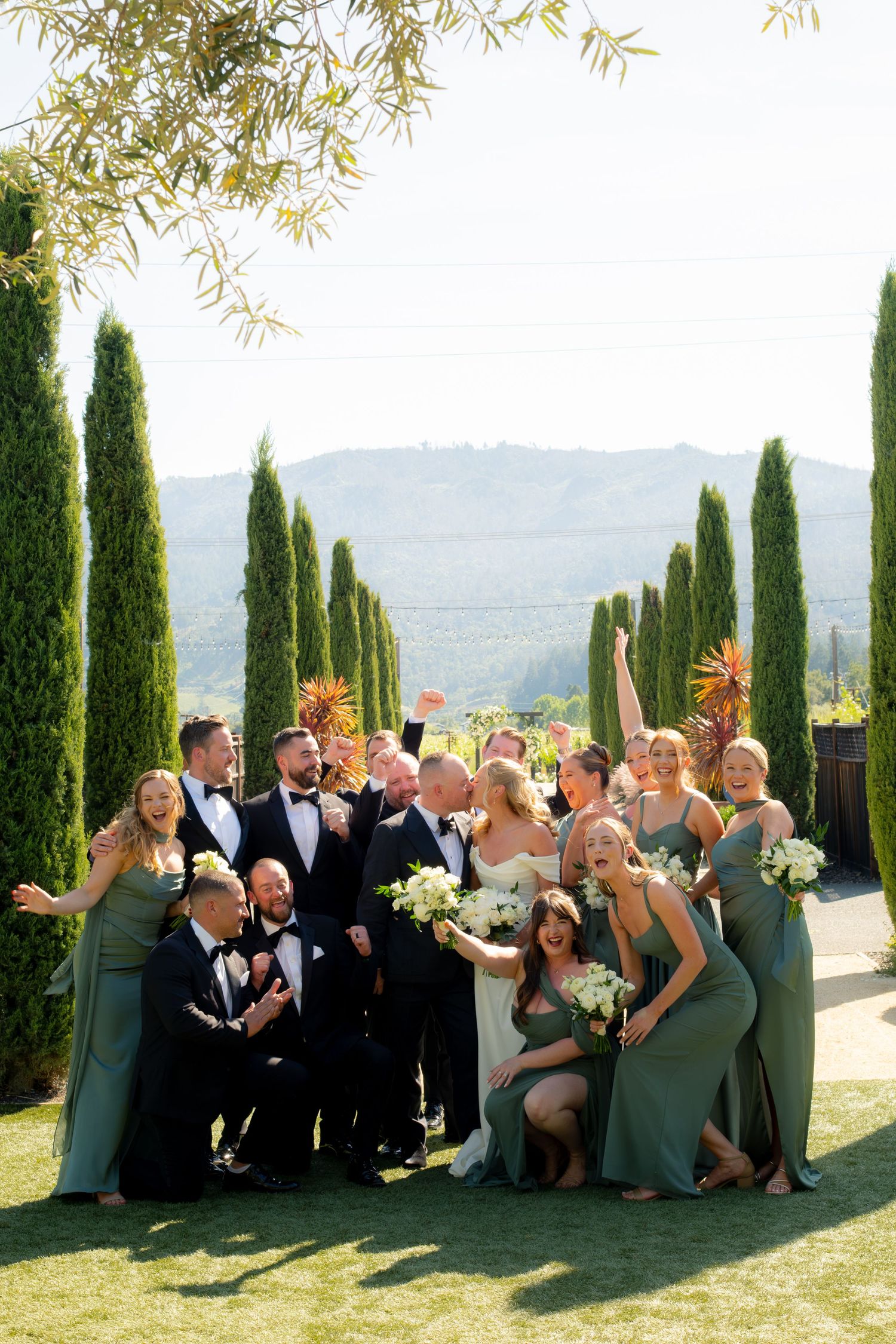 Joyful wedding party in sage green gowns and black tuxedos celebrating with cypress trees and vineyard hills behind them.