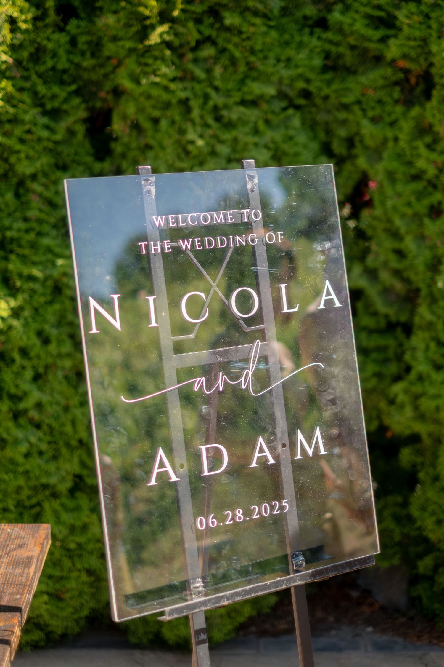 Acrylic wedding welcome sign reading Welcome to the Wedding of Nicola and Adam 06.28.2025 outdoors.