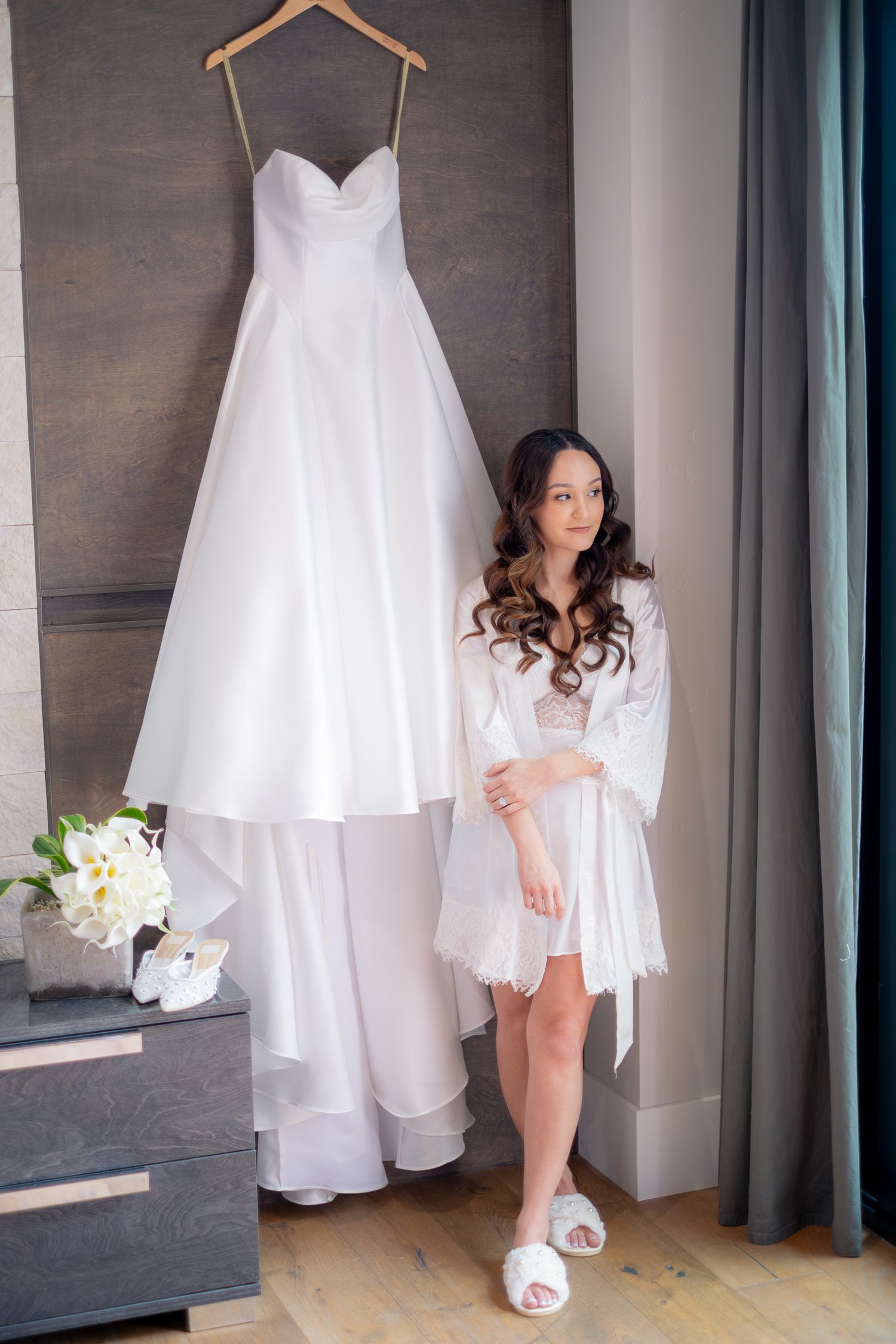 Bride in white lace robe standing next to her elegant A-line wedding dress hanging on a wooden hanger.