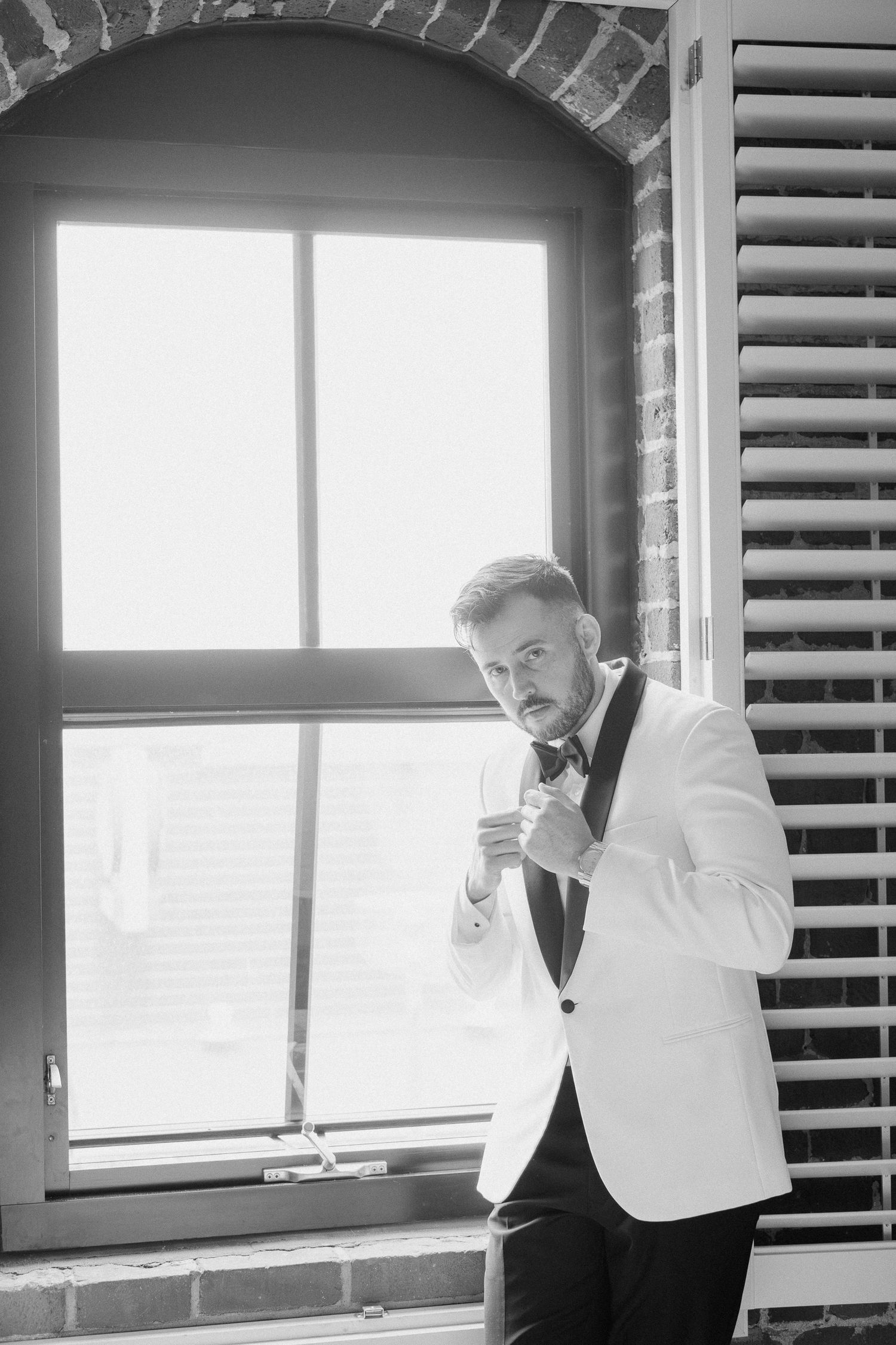 Groom in white tuxedo jacket adjusting bow tie by arched window in black and white wedding photo.