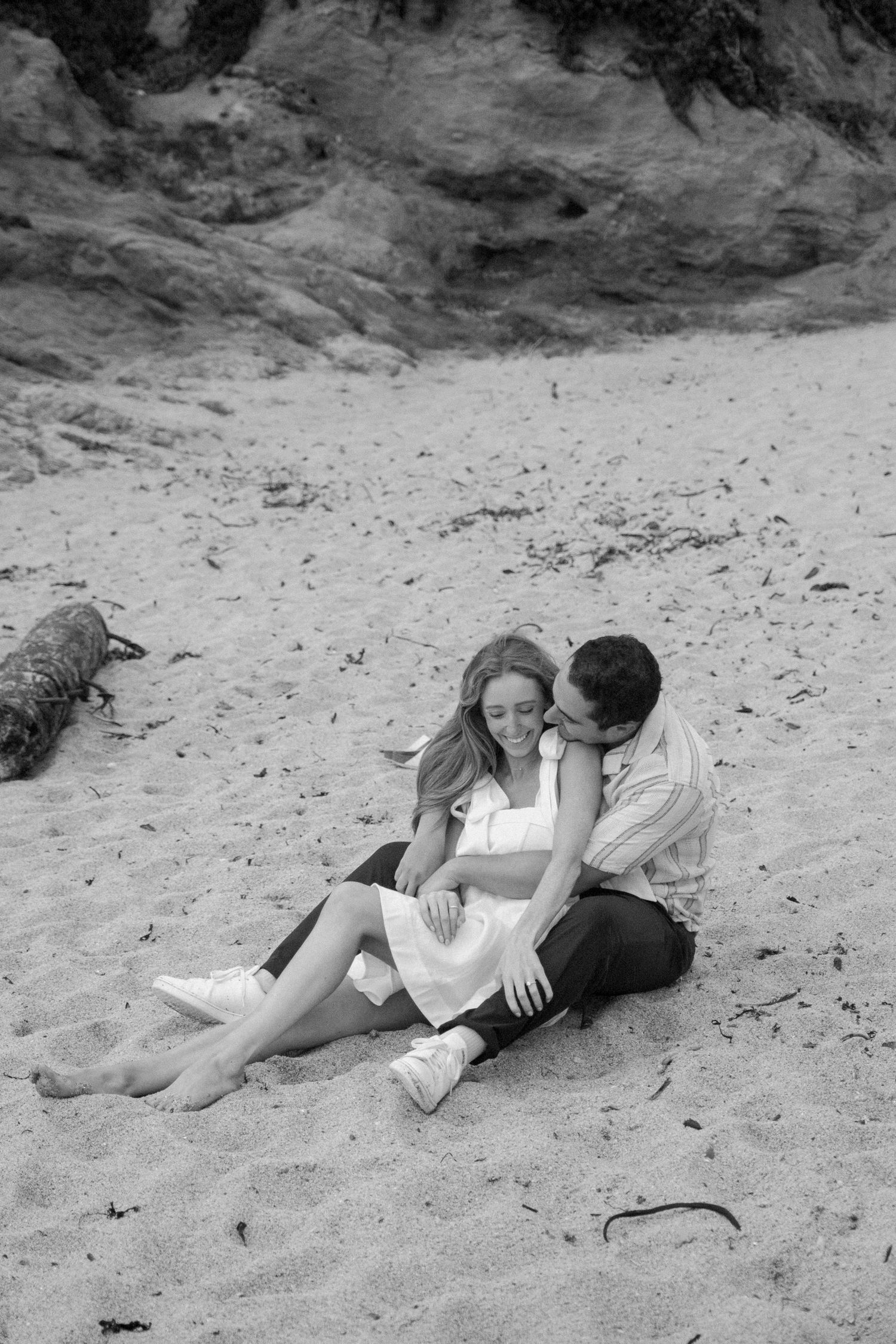 Couple sitting together on sandy beach, man kissing woman's cheek, black and white romantic photography.