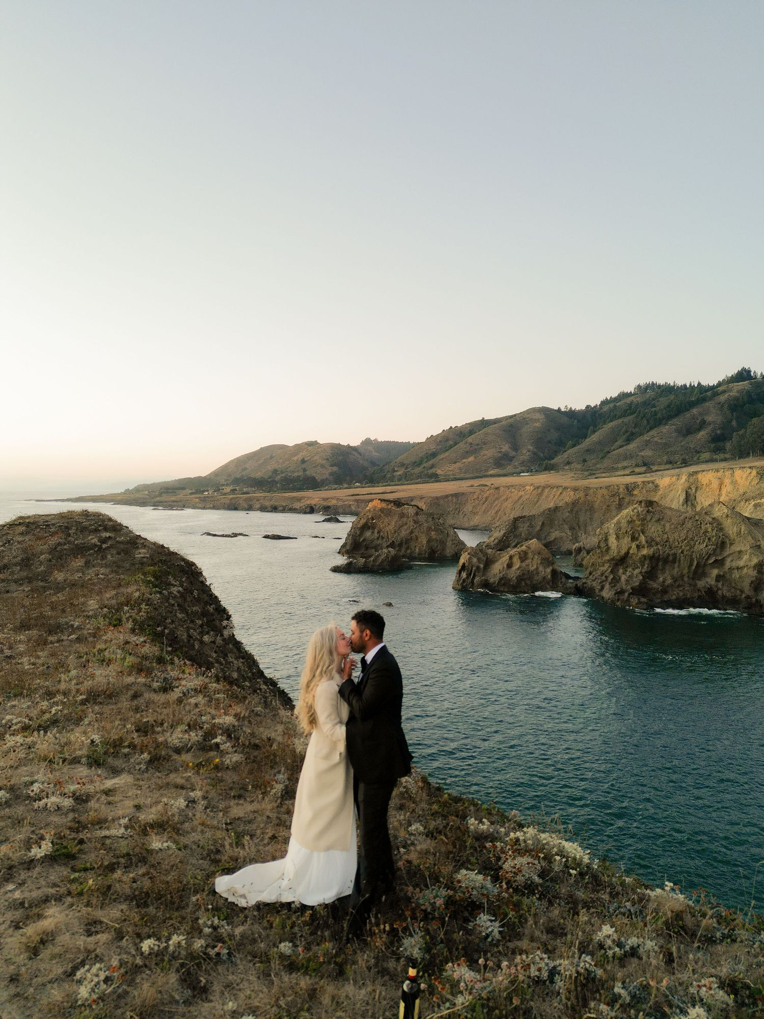 Bride and groom sharing a kiss on dramatic coastal cliffs overlooking a stunning ocean cove.