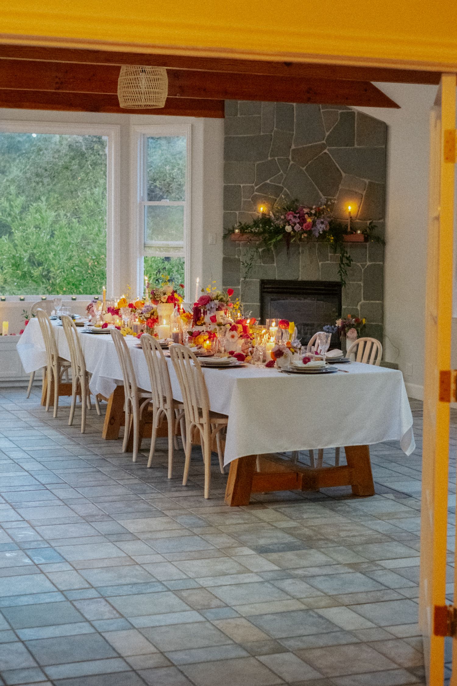 Elegant wedding reception dining room with long table, white linens, colorful floral centerpieces, and stone fireplace.