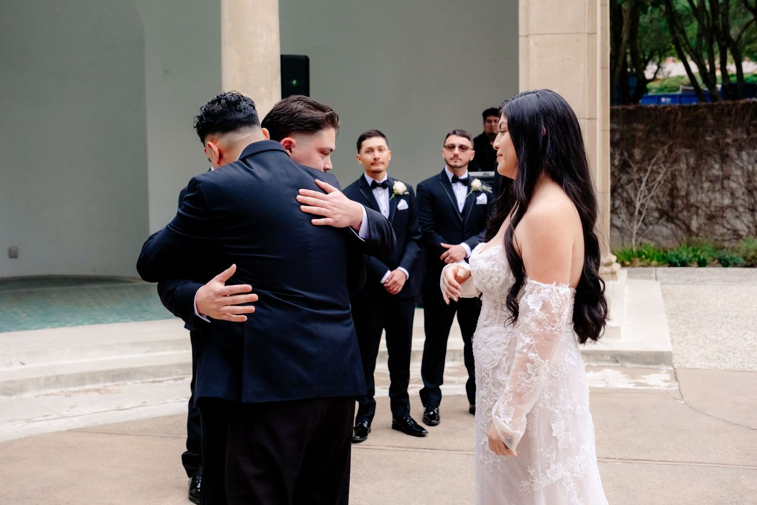 Groom hugging a guest at an outdoor wedding while groomsmen and bride in lace gown look on.