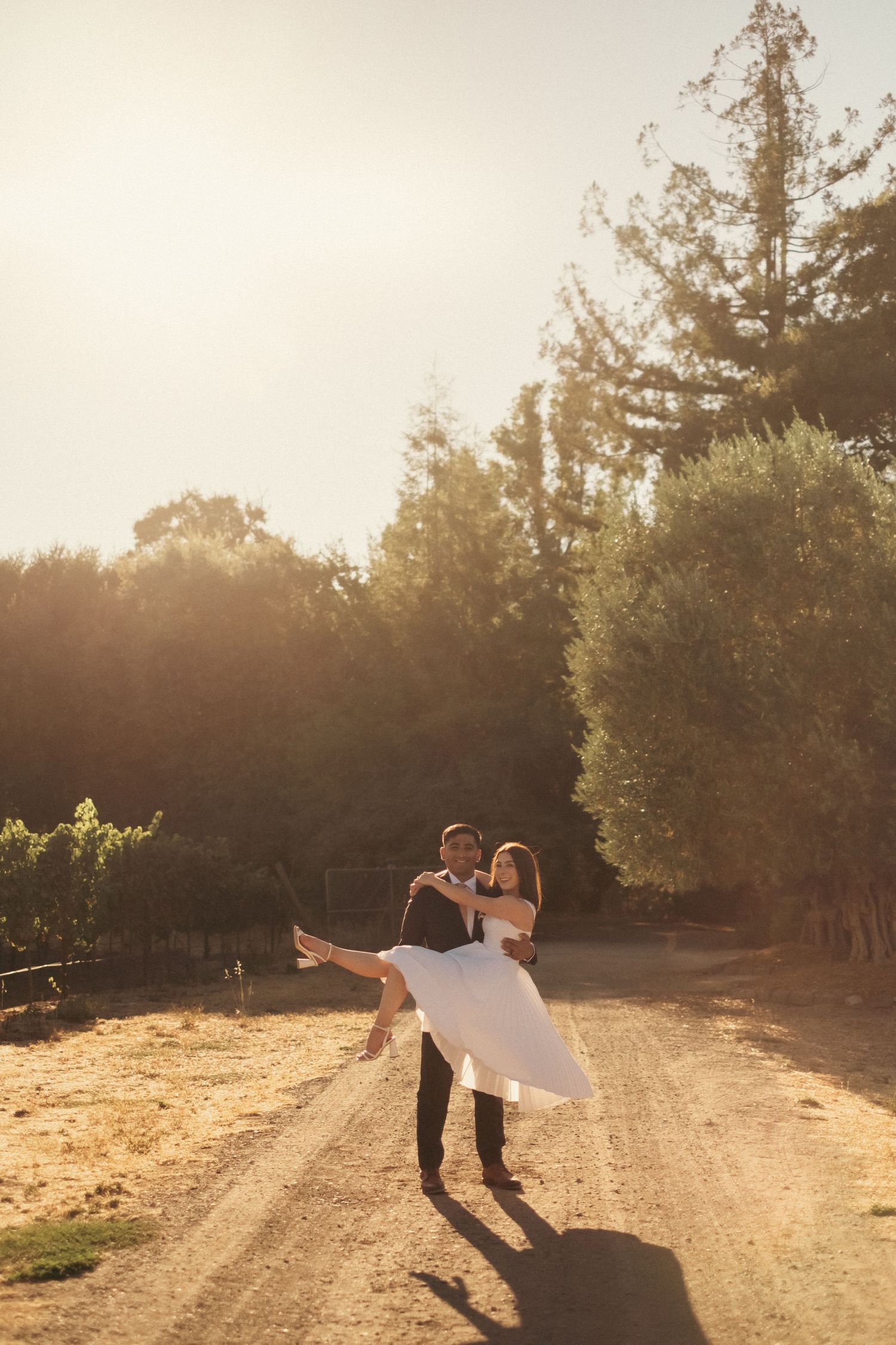 Groom carrying bride in white dress on a sunlit dirt road surrounded by trees and vineyards.