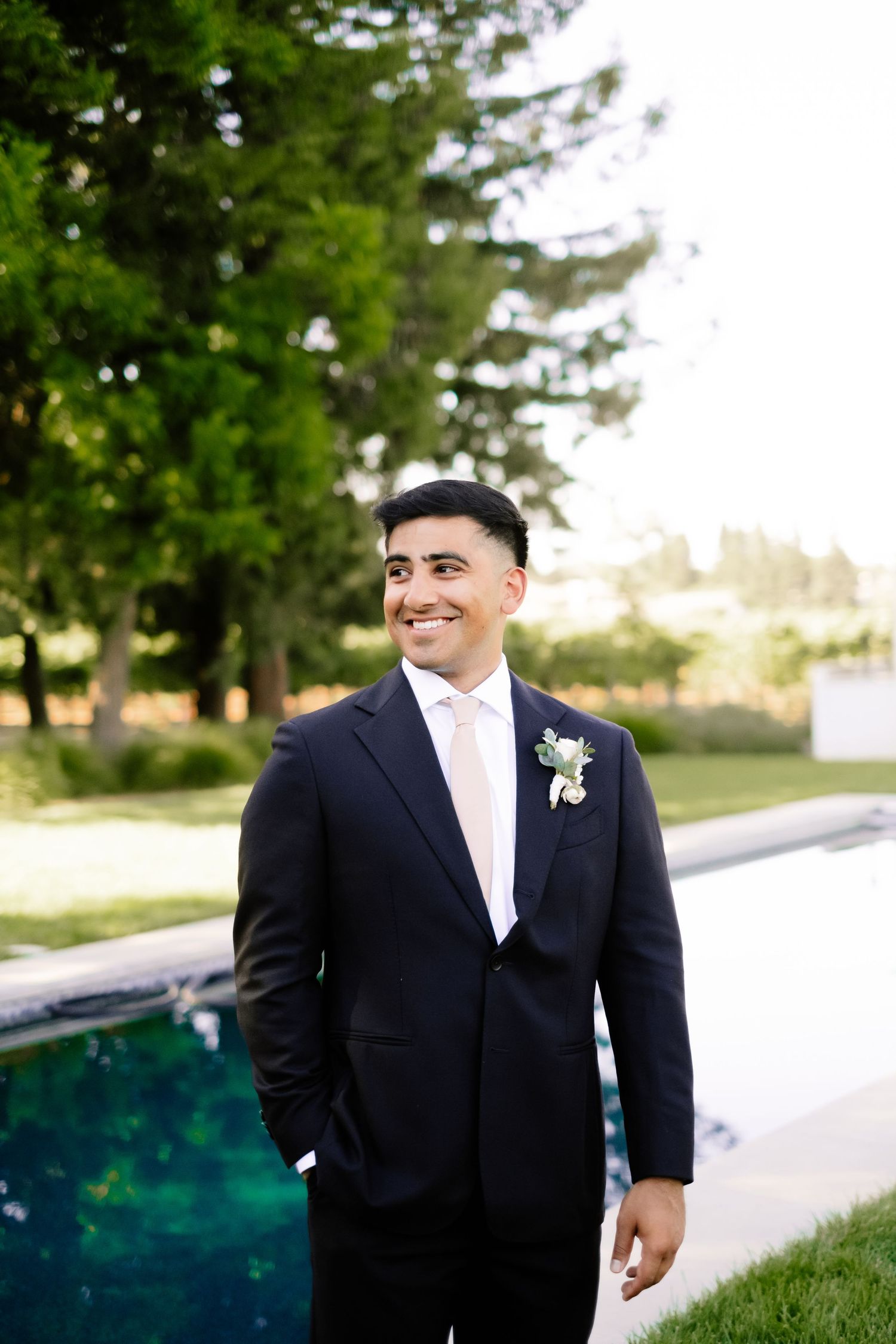 Smiling groom in navy suit with boutonniere standing beside outdoor pool at sunset.