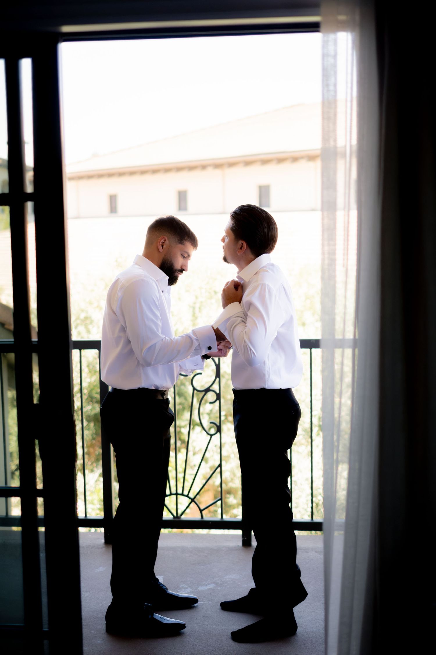 Two groomsmen in white shirts help each other get dressed on a balcony before the wedding ceremony.