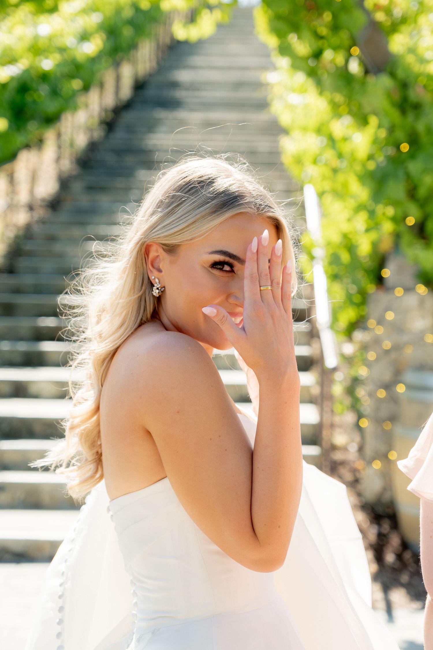 Beautiful blonde bride in strapless white gown poses playfully near vine-covered stone staircase outdoors.