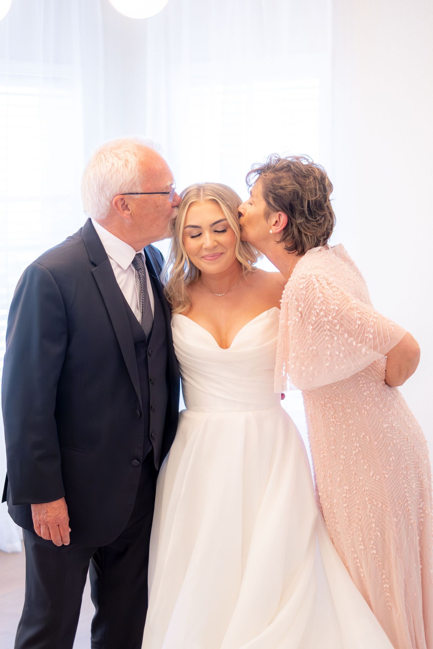 Bride in white wedding gown receives kisses on both cheeks from her proud parents simultaneously.