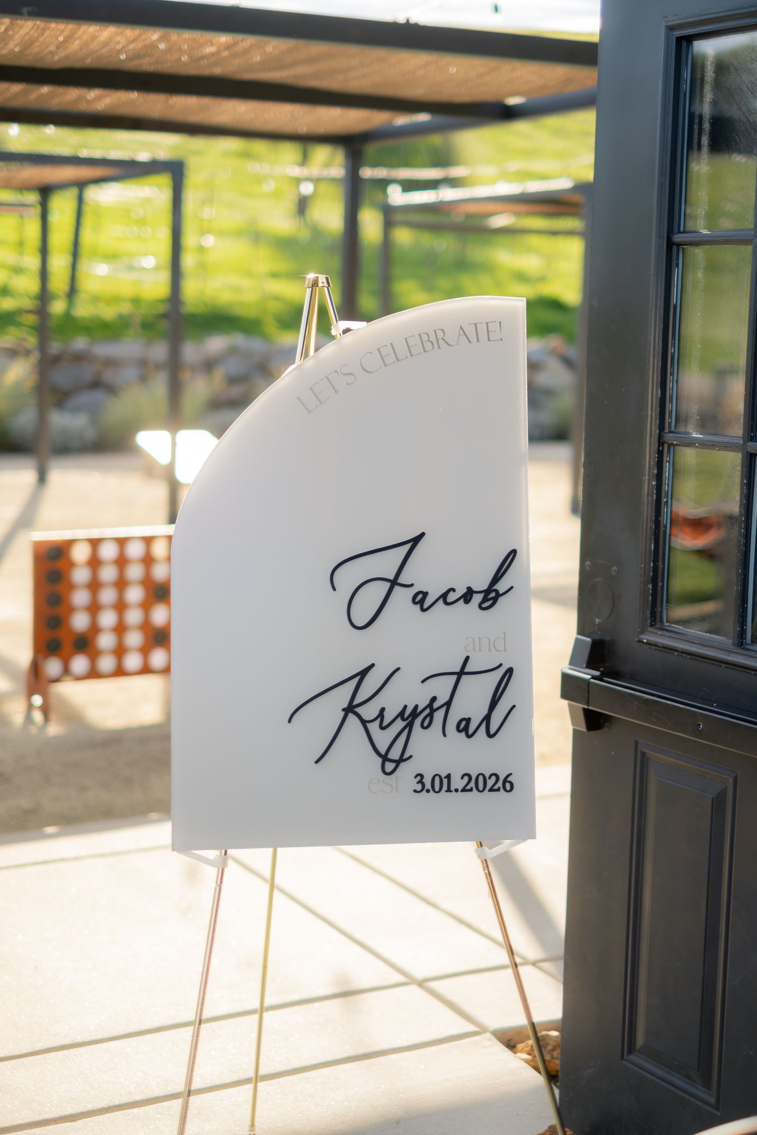 Wedding welcome sign for Jacob and Krystal dated 3.01.2026 displayed on gold easel outdoors.