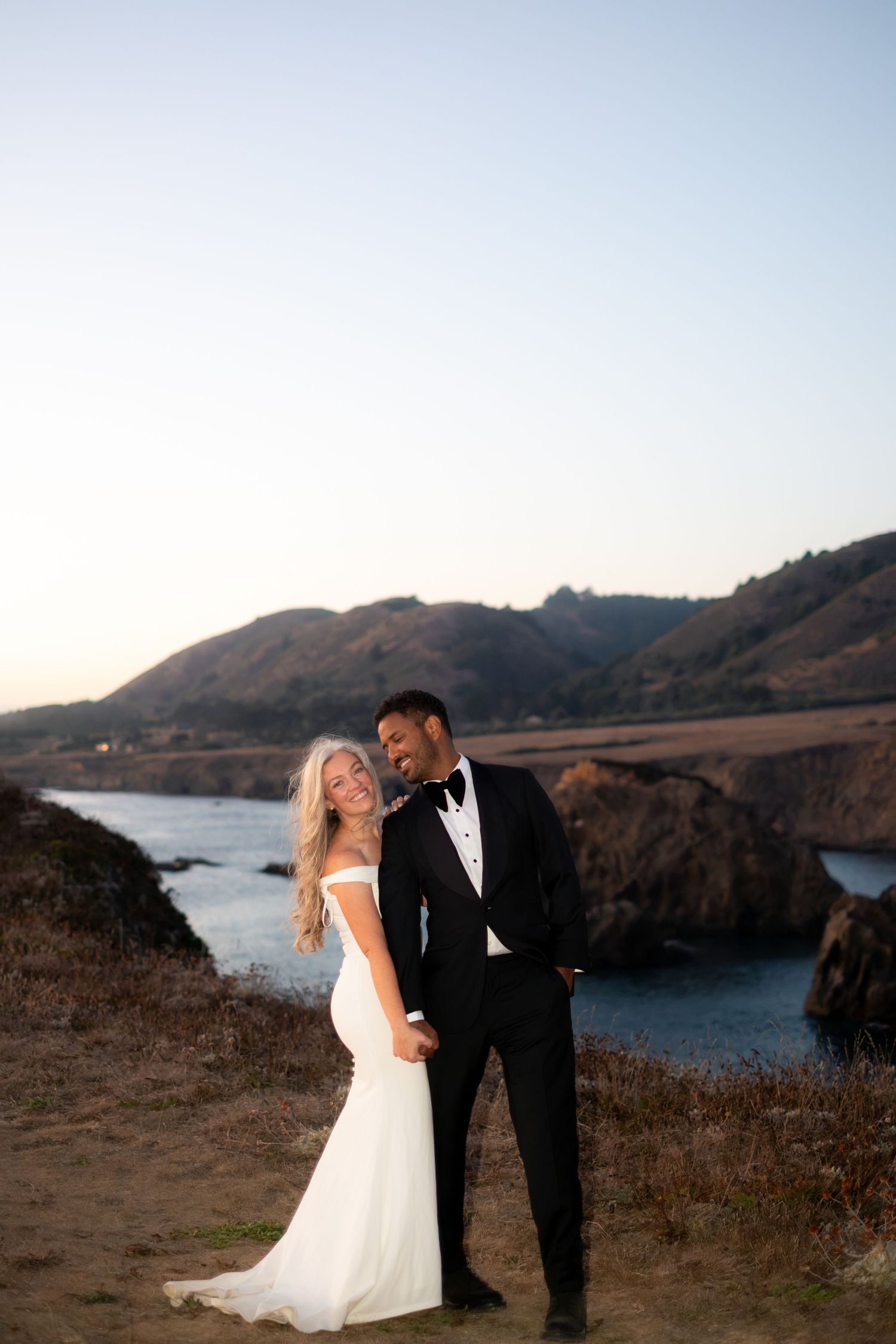 Bride and groom holding hands on a cliffside overlooking a stunning coastal bay at sunset.