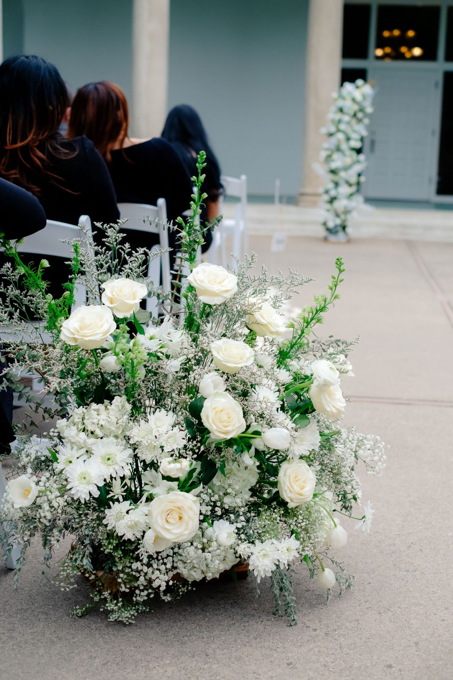 Lush white roses and baby's breath floral arrangement decorating a wedding ceremony aisle outdoors.