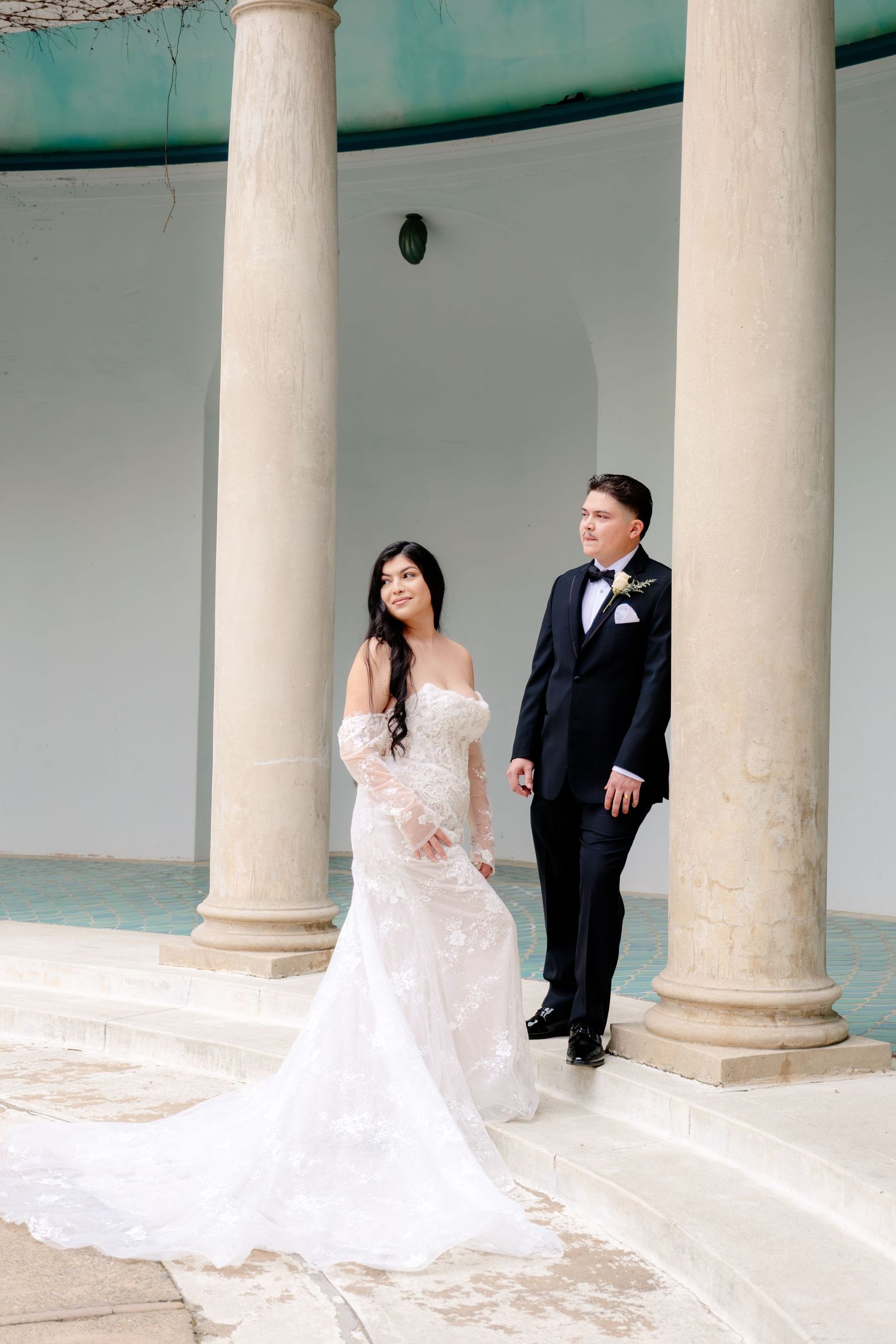 Bride in fitted lace gown and groom in black tuxedo pose between white columns at wedding.