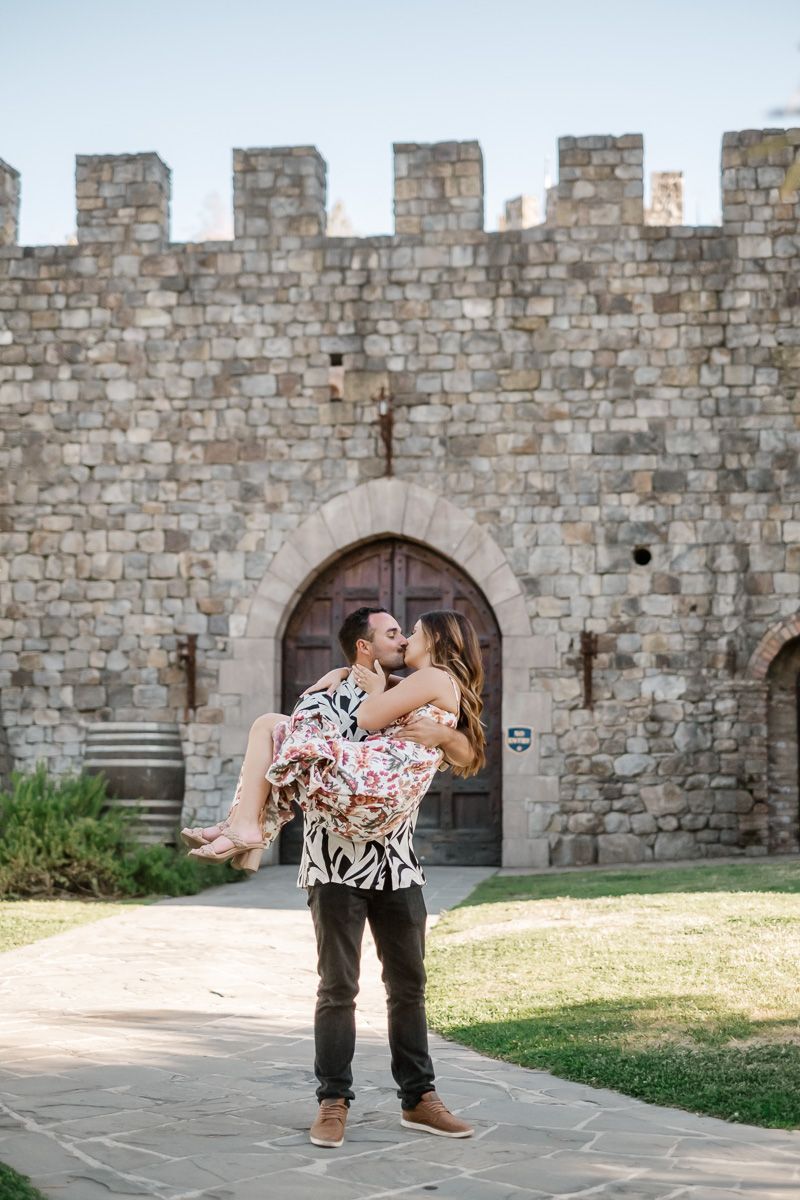 Couple kissing in front of a stone castle entrance, man lifting woman in floral dress at a winery.