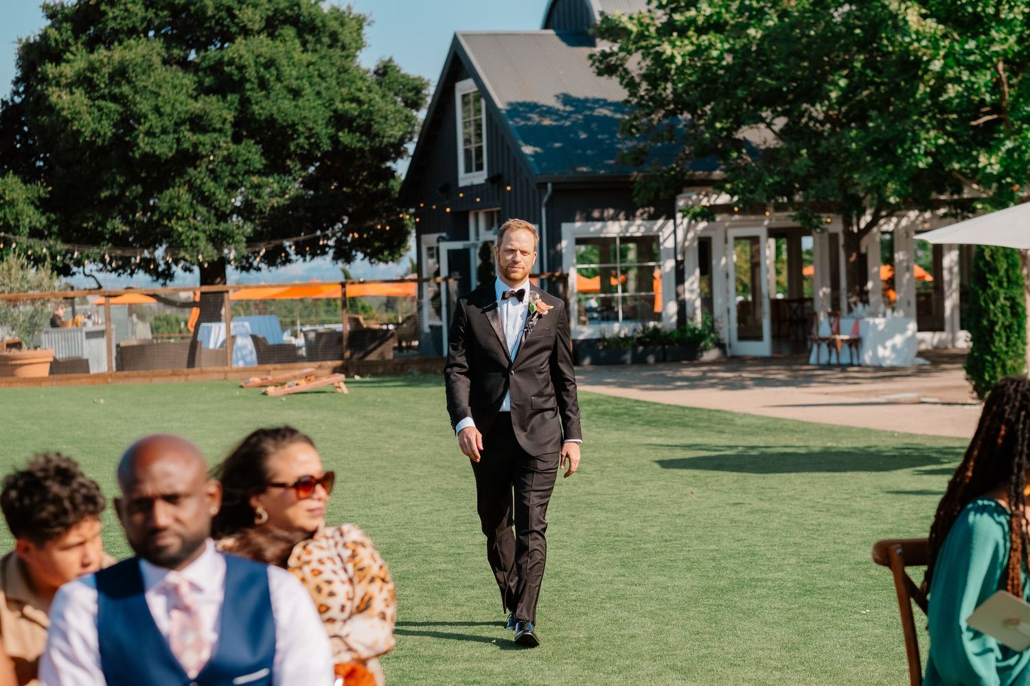 Groom in a dark suit walks down the aisle at an outdoor wedding ceremony on a green lawn.