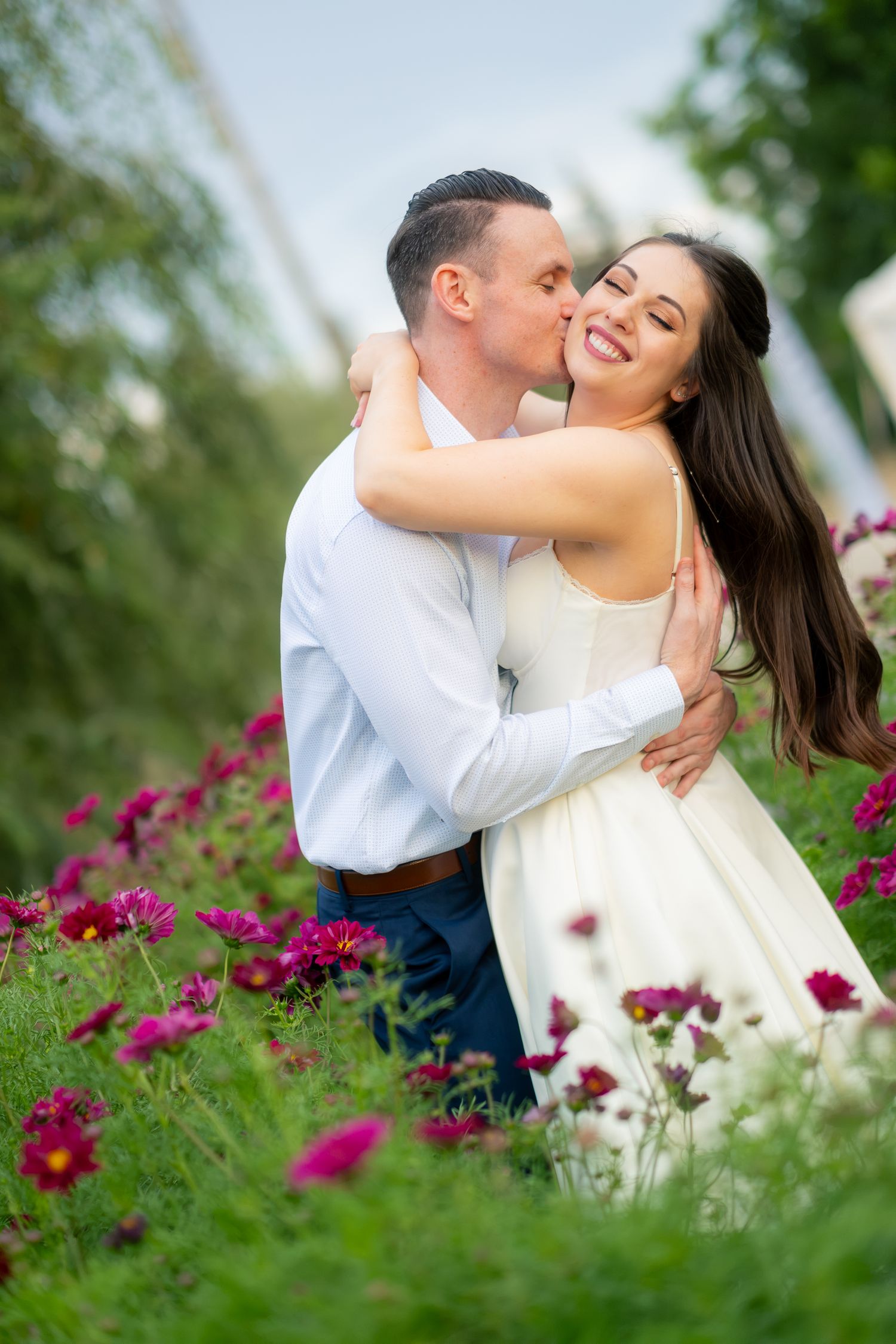 Groom kissing smiling bride on the cheek among vibrant pink flowers outdoors.