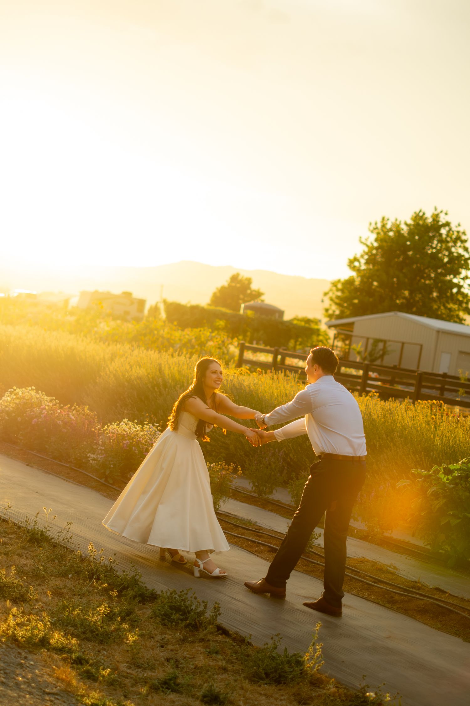 Bride and groom dancing hand in hand on a sunlit path at golden hour with lavender fields and mountains in background.