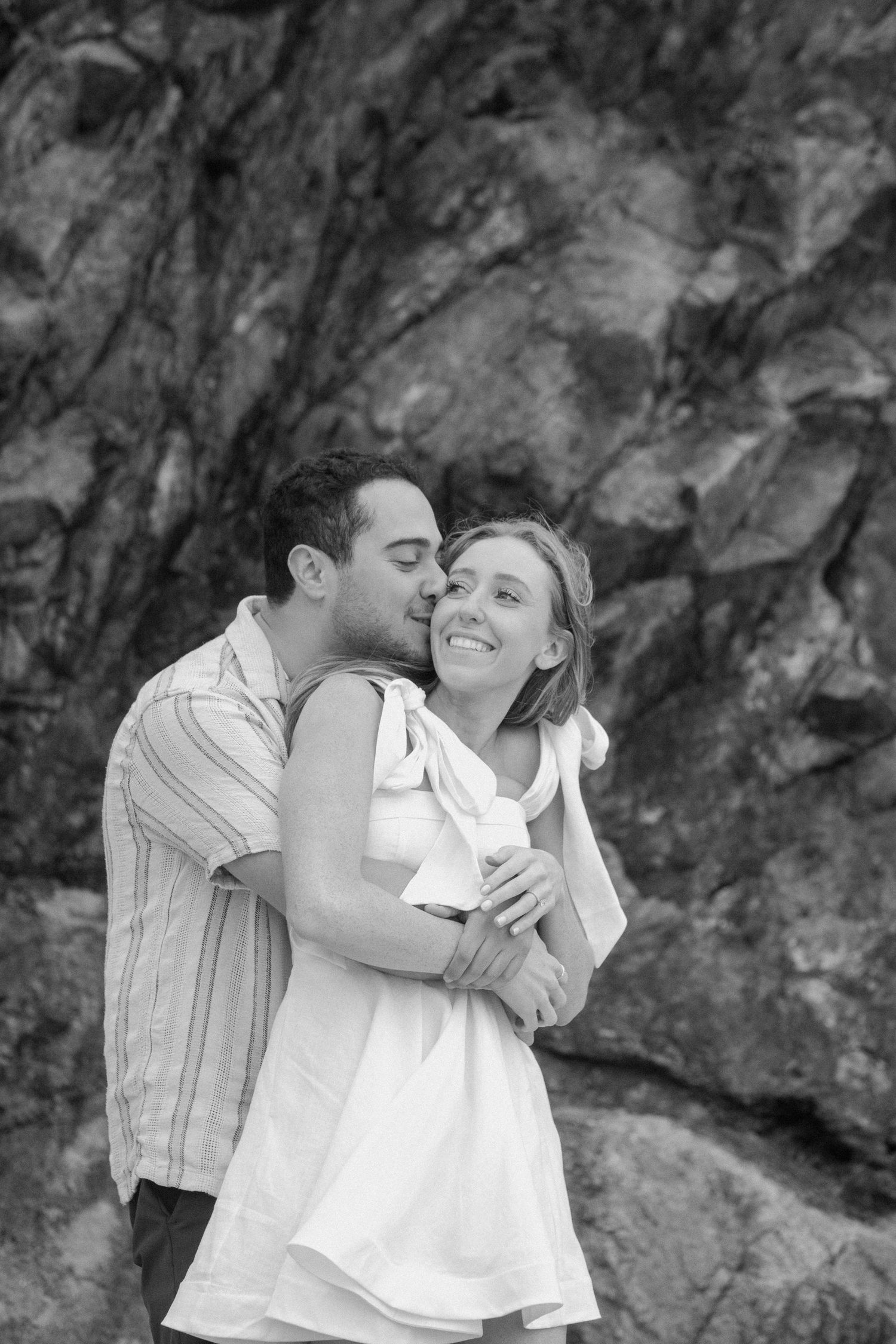 A man kisses a smiling woman on the cheek against a rocky backdrop in black and white.