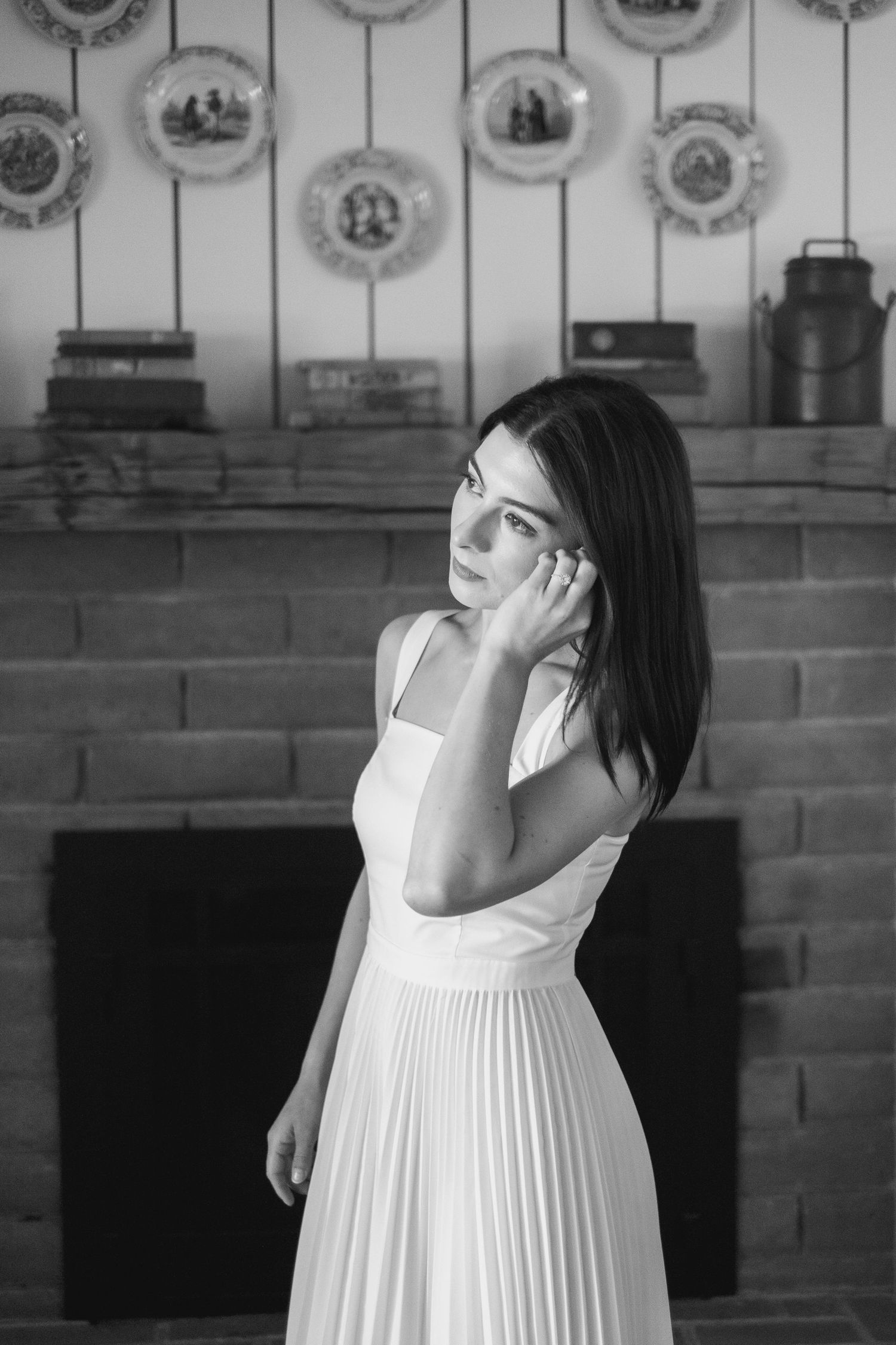 Black and white portrait of bride in white pleated dress standing by stone fireplace.