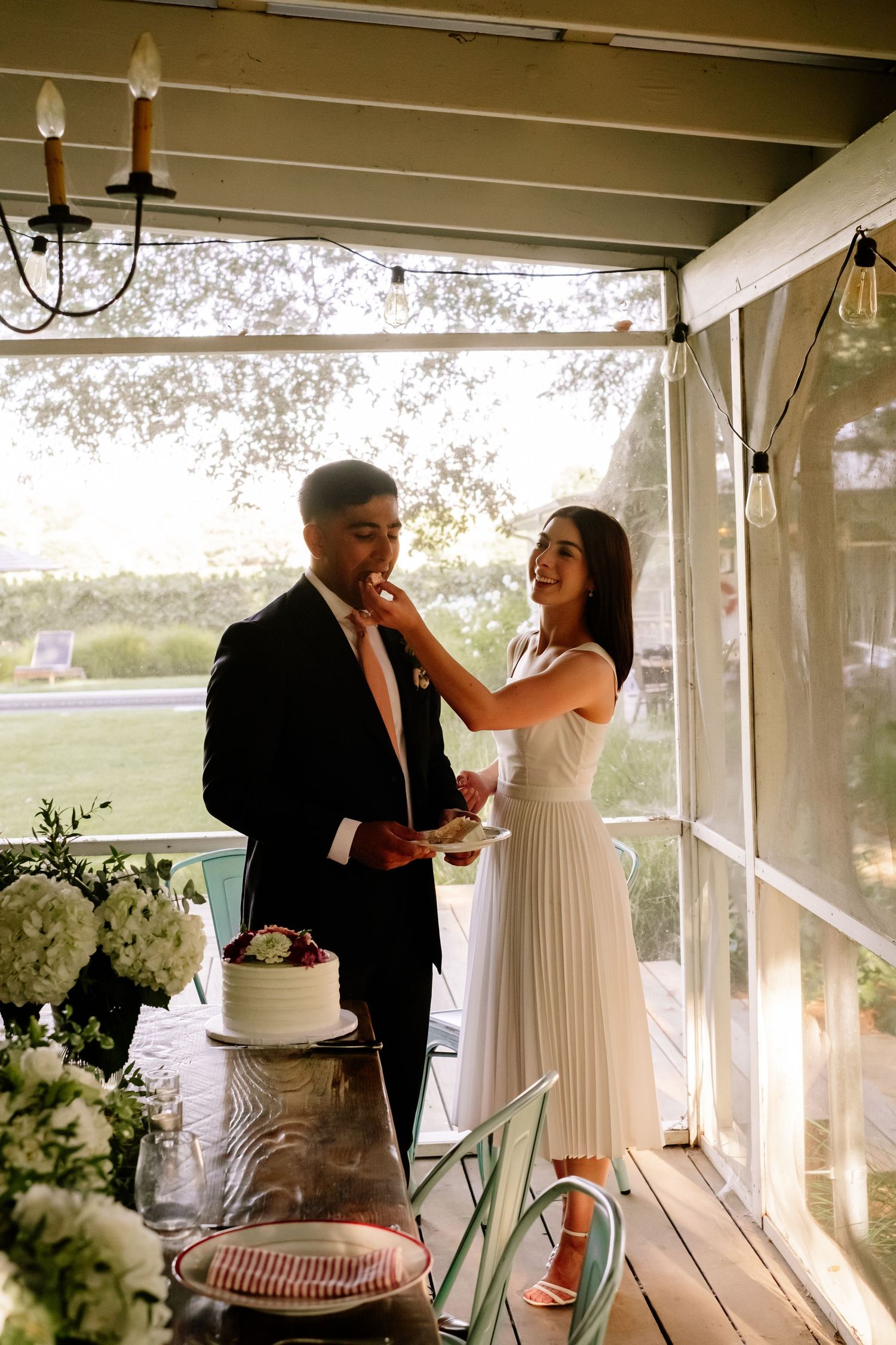 Bride feeds groom wedding cake on decorated porch with white hydrangea arrangements nearby.