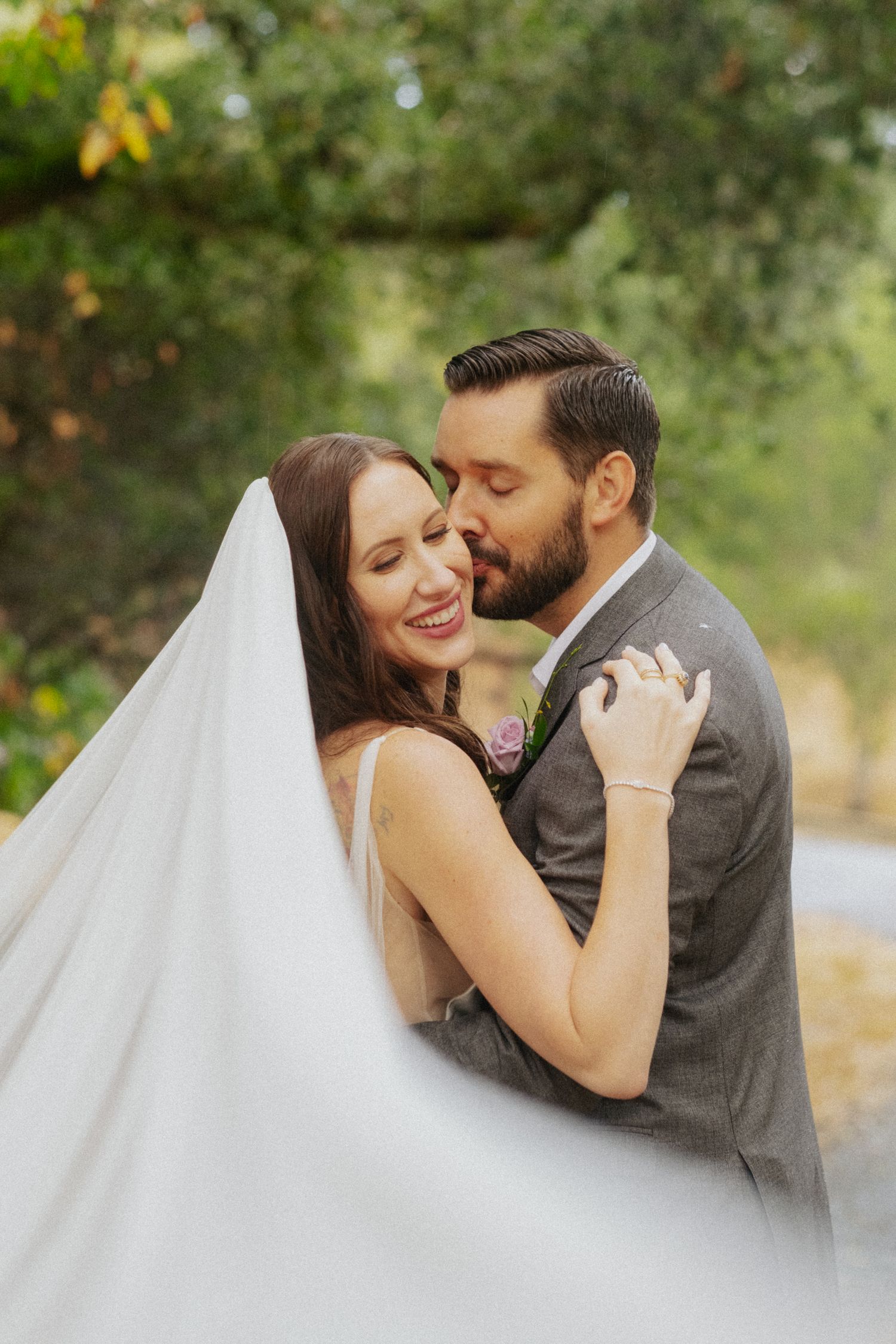 Groom kisses bride on cheek as her veil flows in outdoor wedding portrait.