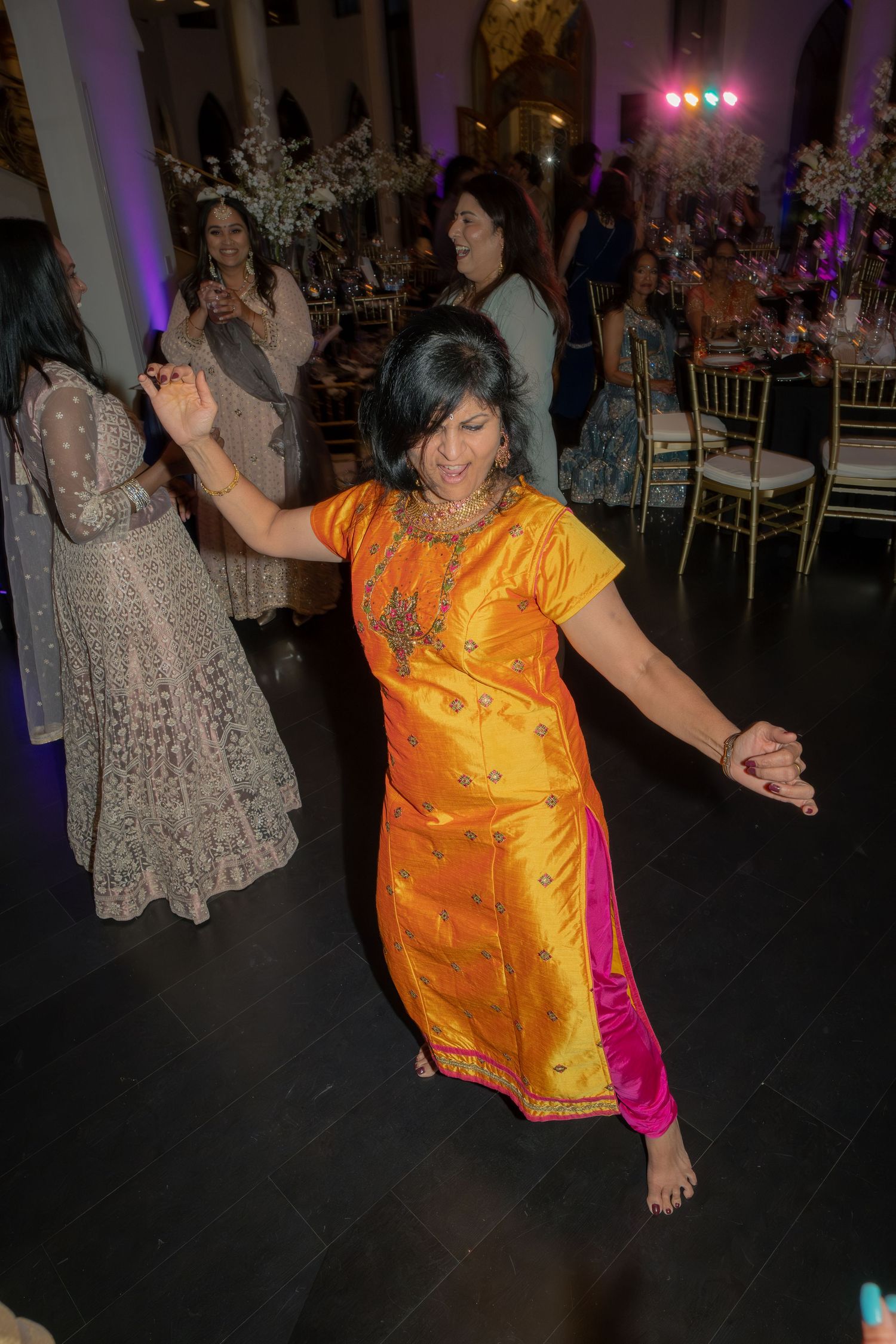 Woman dancing barefoot in orange and pink Indian salwar kameez at a vibrant wedding reception.