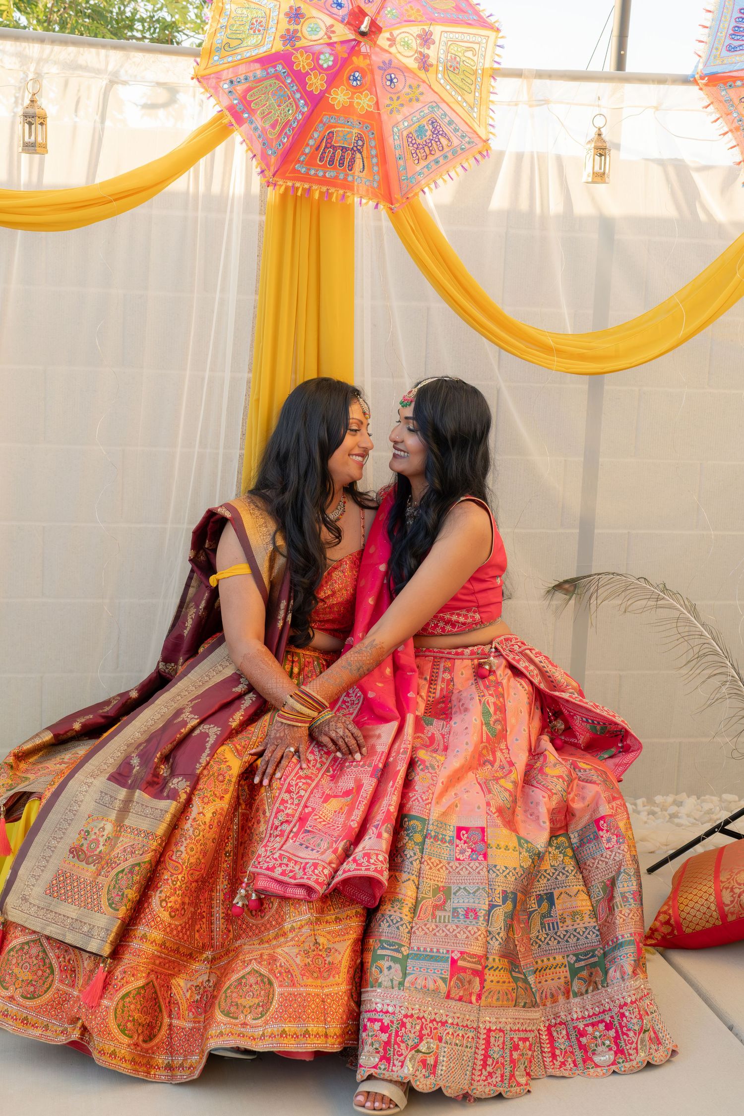 Two women in vibrant traditional Indian lehengas share a joyful moment at a colorful wedding celebration.