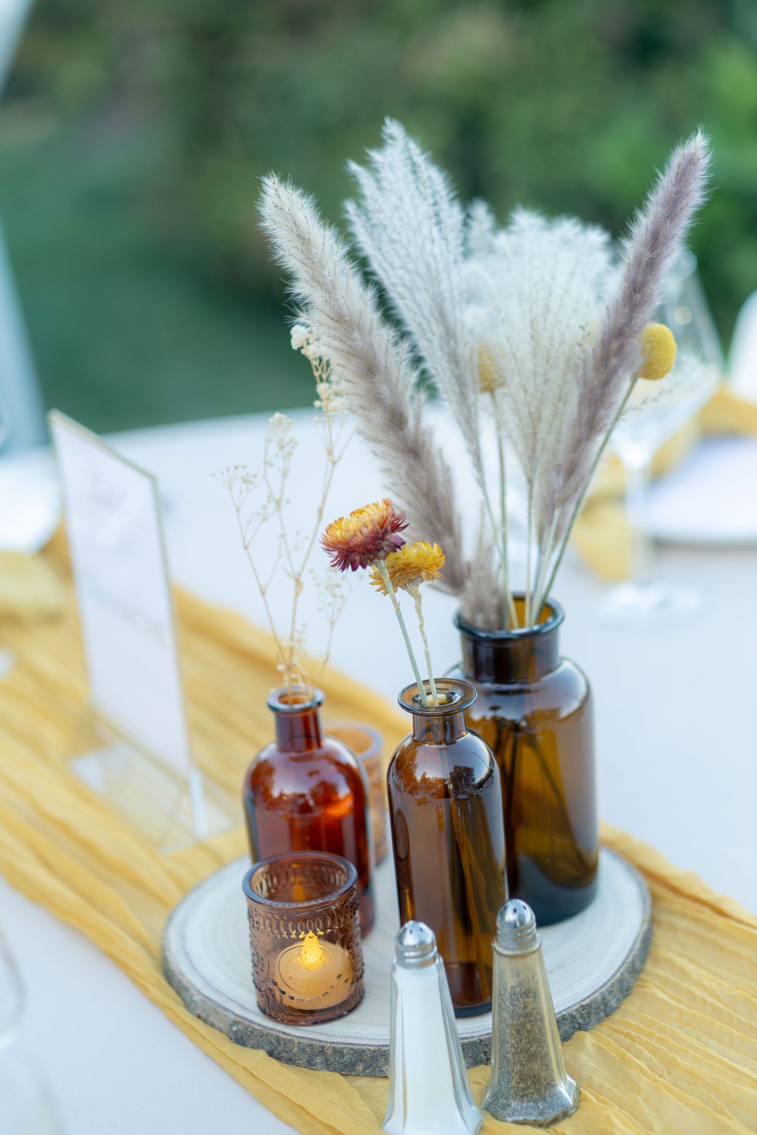 Rustic wedding table centerpiece with dried pampas grass in amber glass bottles on a wood slice with a candle.