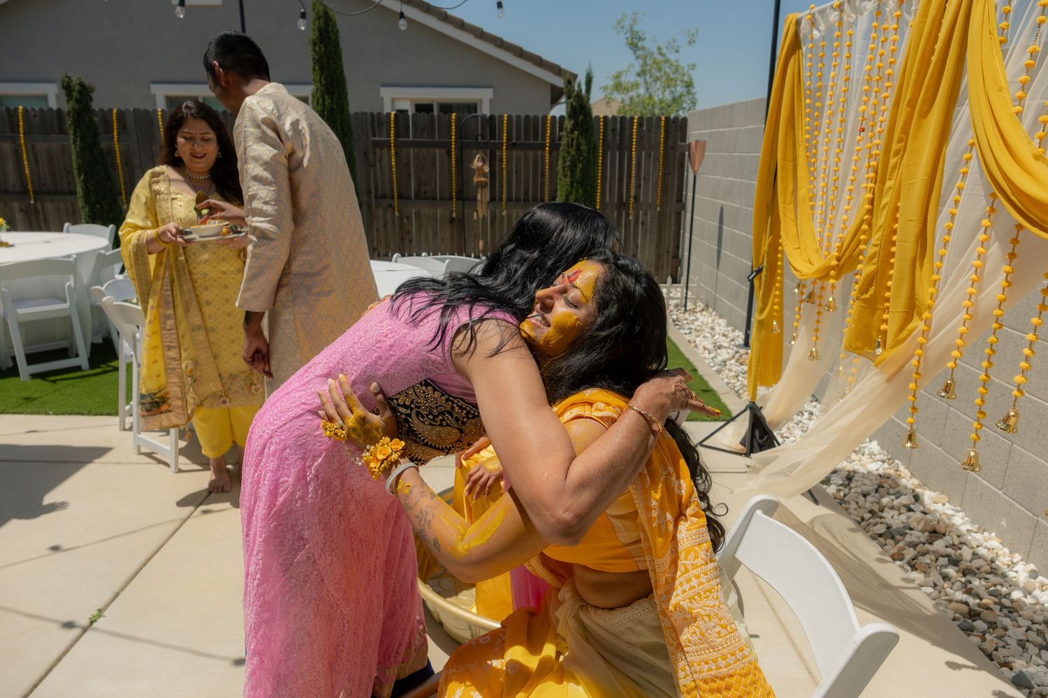 Two women in vibrant yellow and pink Indian sarees share a warm embrace at an outdoor South Asian wedding celebration.