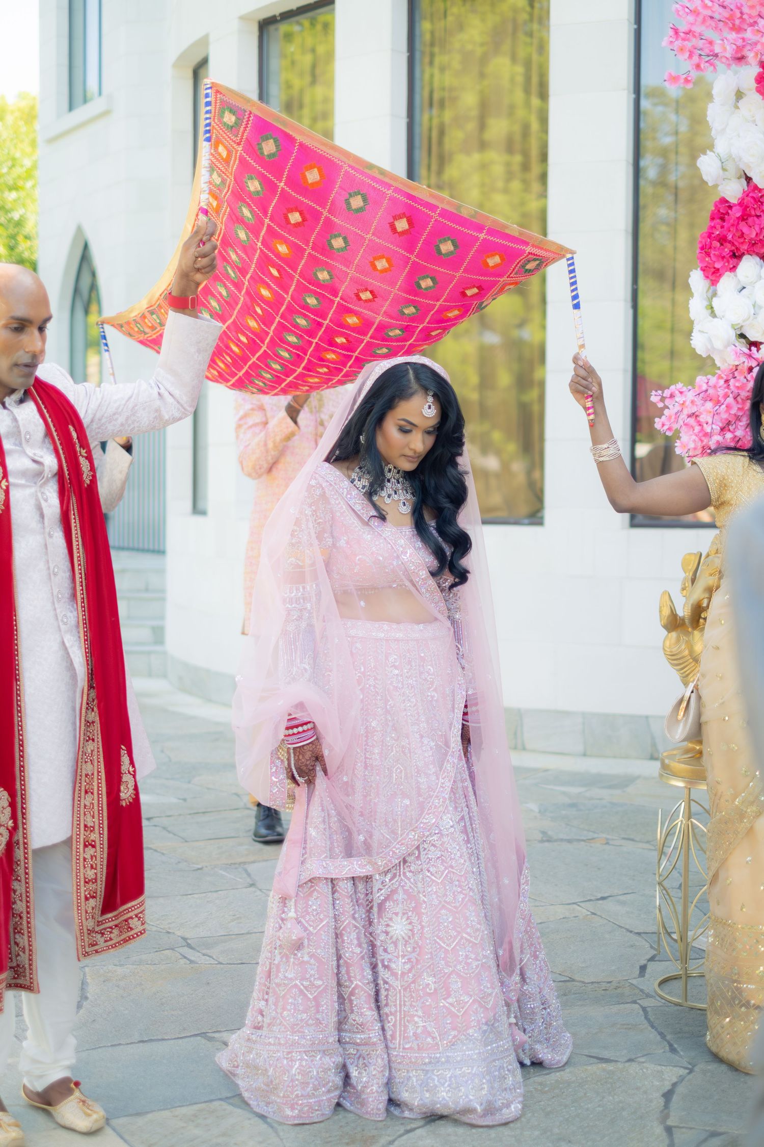 Indian bride in pink lehenga walking under pink chunni during traditional wedding entrance ceremony.